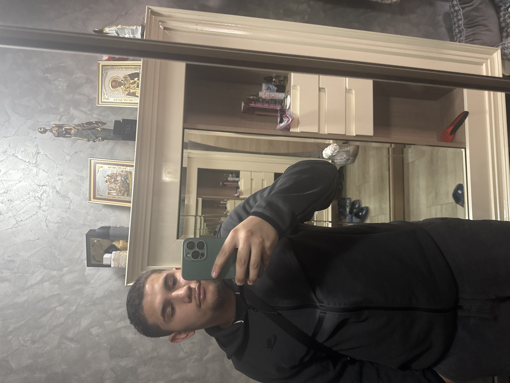 A man is taking a selfie in front of a large mirror. The mirror is framed with a beige wooden frame. The reflection shows shelves filled with various items, including icons, a figurine, toiletries, and shoes. The man is wearing a gray hoodie with a black strap across his chest. The background wall is textured with a gray and white pattern. There is no indication of the city of Sofia, Bulgaria, in the image.