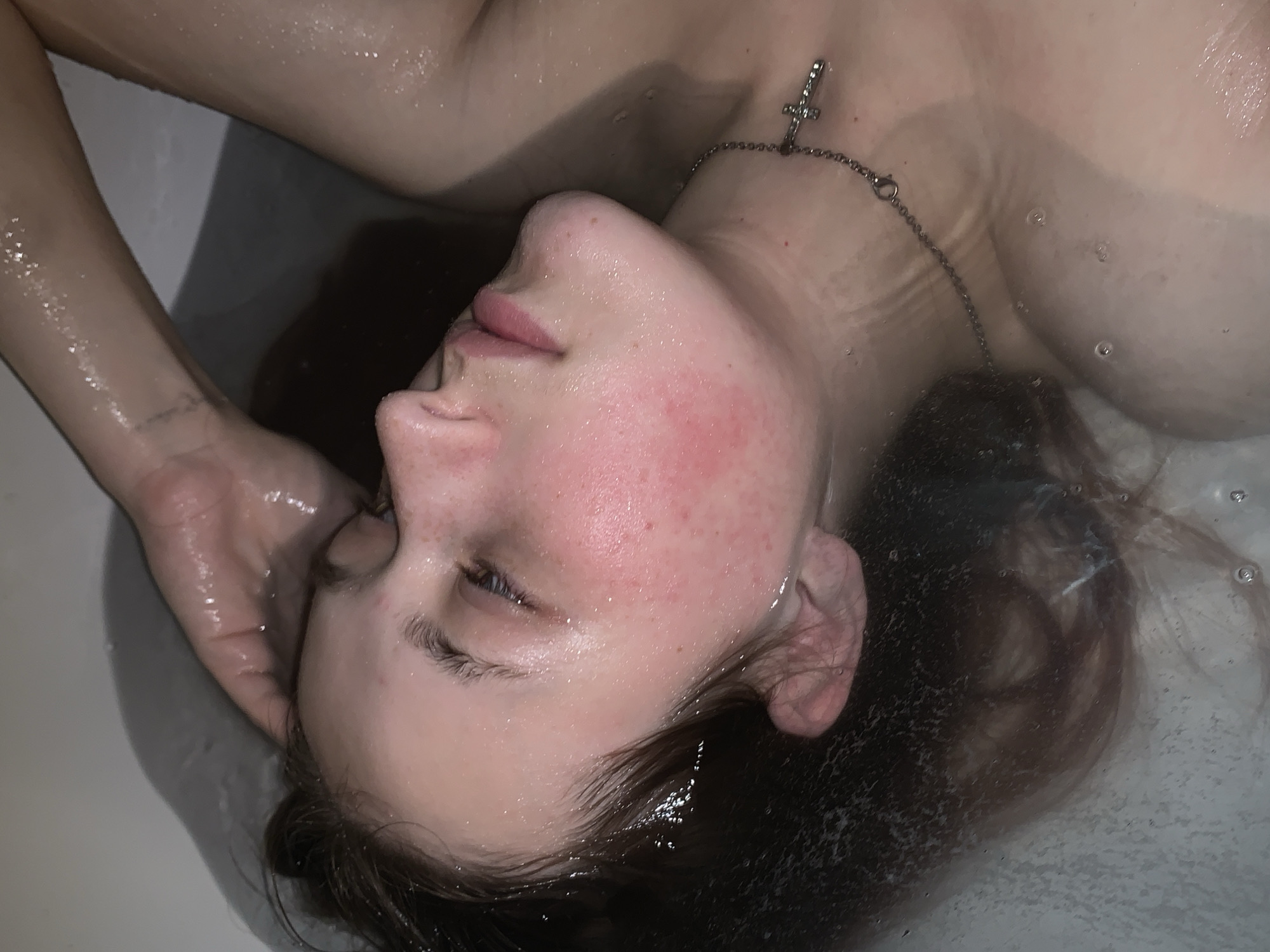 A person is lying in a bathtub filled with water. The individual appears to be submerged up to their neck, with their head tilted back and eyes partially closed. A silver necklace with a cross pendant is visible around their neck. The person's left hand rests on their forehead. The environment appears to be a bathroom, with the bathtub being the primary focus. The location Фоки, Russia, cannot be determined from the image.