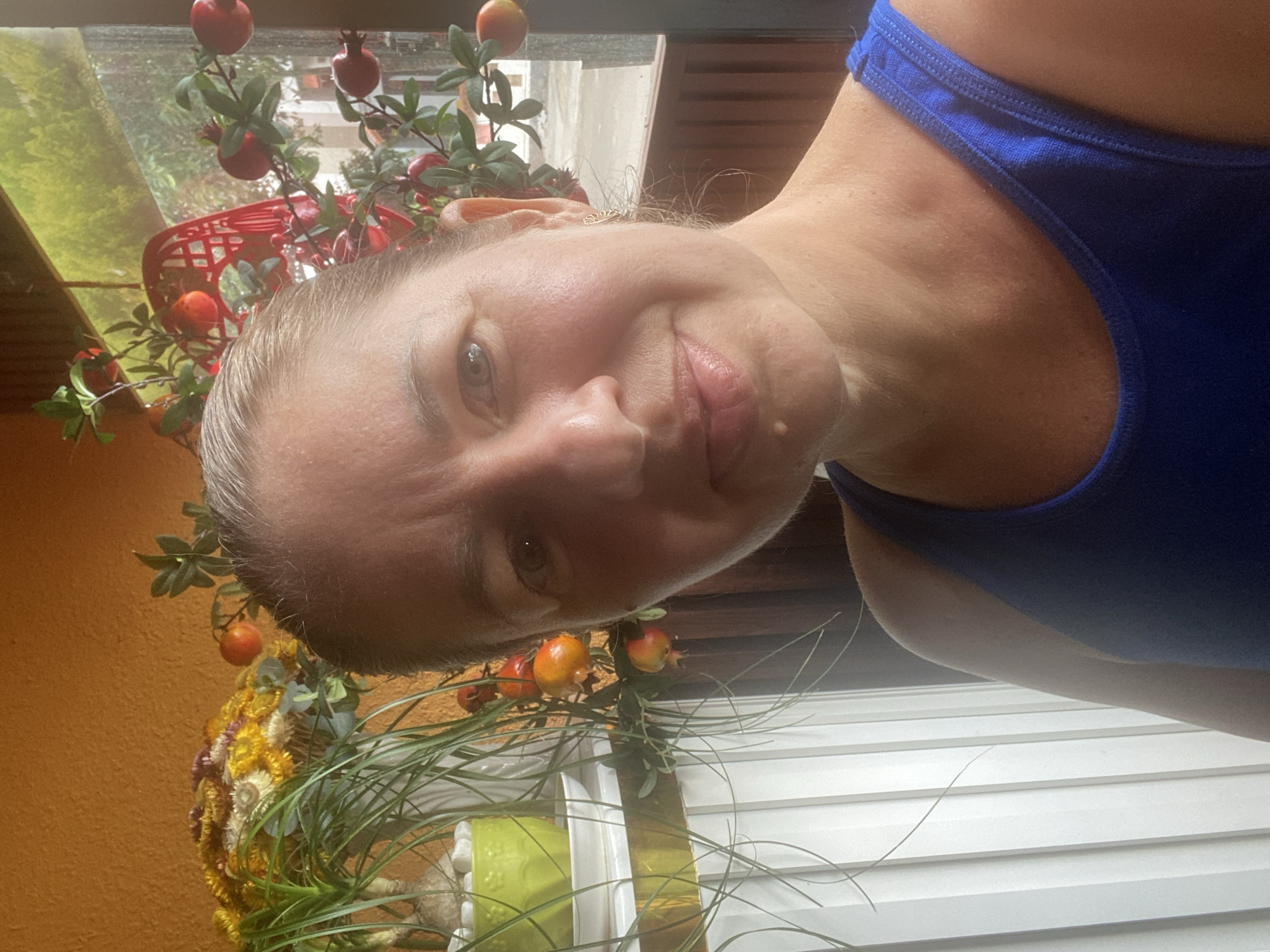 The image is a close-up selfie taken by a middle-aged woman with light skin, graying blonde hair pulled back, and light-colored eyes. She is wearing a blue tank top. The background suggests an outdoor or semi-outdoor setting, possibly a balcony or veranda, with a textured orange wall on the left. To the left of the woman is a plant in a bright green pot with long, green leaves and a bouquet of dried yellow and purple flowers. To the right and behind her, there is a decorative string of artificial pomegranates and leaves, with a red basket hanging in the background. Sunlight is illuminating her face from the right side. The overall scene appears to be in Mũi Né, Vietnam, given the context provided.