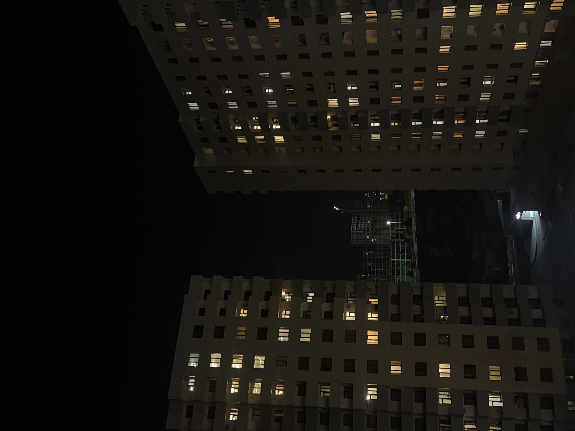 The image is a night photograph featuring two tall, multi-story buildings, oriented horizontally at the top and bottom of the frame, separated by a wide expanse of dark sky. The buildings have a grid-like facade with numerous windows. Many windows are illuminated, casting warm yellow and cool white light, indicating occupancy, while others remain dark. Architectural details like recessed sections or balconies are visible, particularly on the edges of the buildings. On the building occupying the bottom portion of the frame, external air conditioning units are discernable beneath some windows. Faint outlines of other structures or scaffolding can be seen in the dark space between the two main buildings, towards the right. The dark background confirms it is nighttime with a clear sky. No people or visible text are present. The scene depicts an urban environment, likely in Yerevan, Armenia.