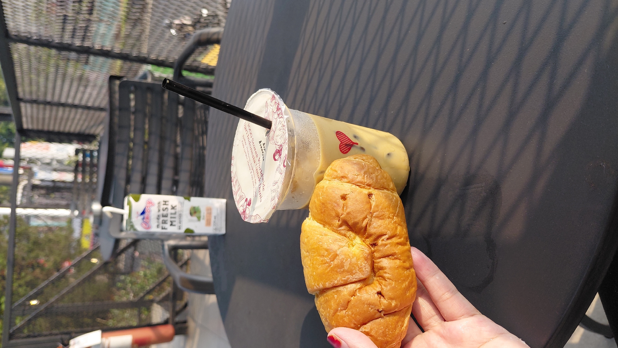 A hand with red-painted fingernails holds a golden-brown croissant on a dark-colored outdoor table. To the right of the croissant is a clear plastic cup filled with a light-colored iced beverage, topped with a sealed lid and a black straw. A small red heart logo is visible on the side of the cup. Behind the cup, a small carton of "Cimory FRESH MILK" stands upright. The table is casting long, diagonal shadows from an overhead structure. In the background, a metal mesh fence encloses an outdoor seating area with dark chairs. The setting appears to be an outdoor cafe in the city of Karanglewas, Indonesia.