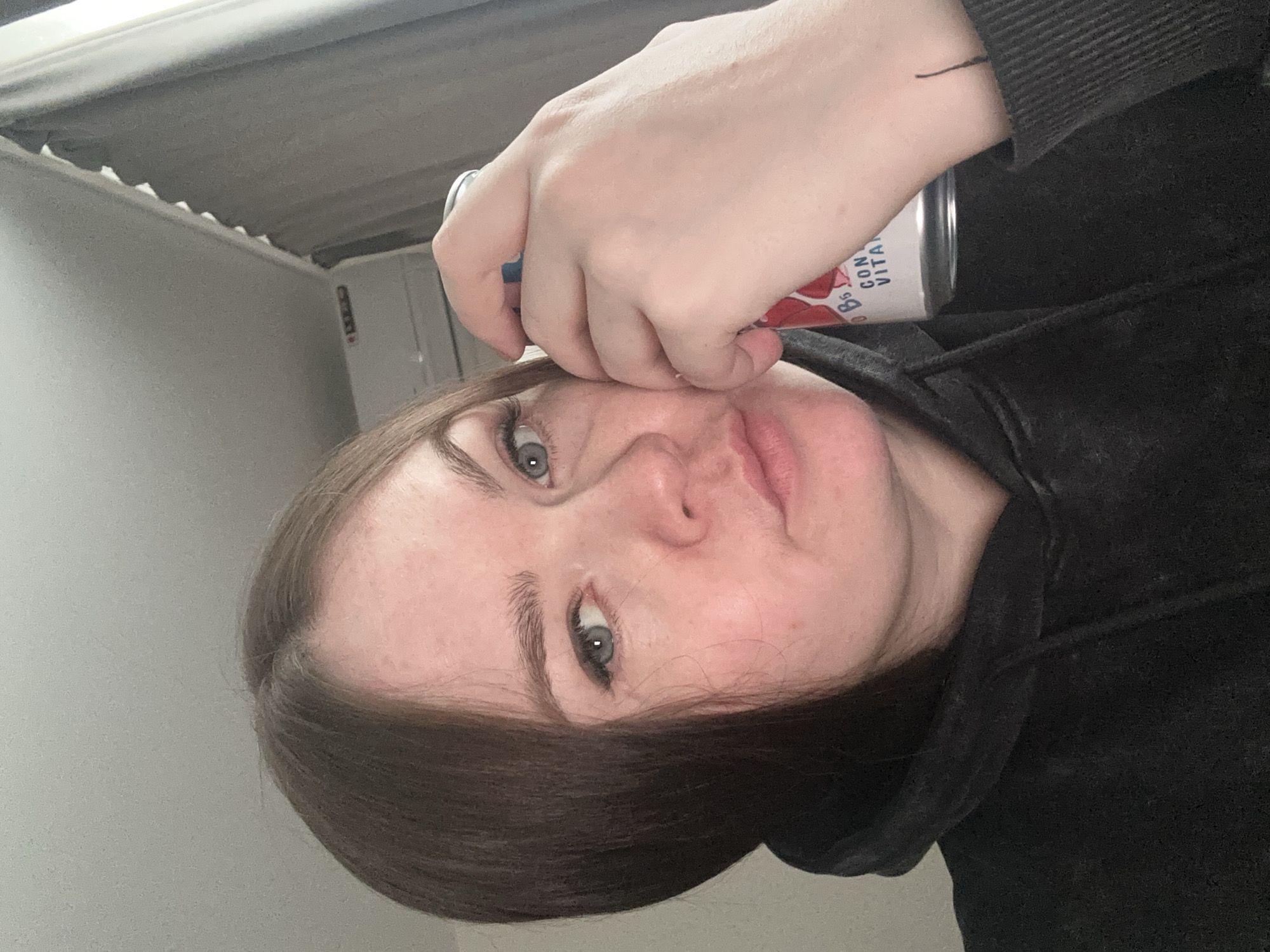 The image is a close-up, rotated portrait of a person, likely a selfie. The subject has fair skin, dark brown hair parted on the side, and light-colored eyes, appearing blue or grey, which are directed at the camera with a neutral expression.
The person's left hand holds a red and white can against their forehead, resting just above their right eyebrow. Visible text on the can includes "B6" and "CON VITA". A thin, dark, linear tattoo or mark is visible on the inner wrist of the hand holding the can. The person is wearing a dark, possibly black, long-sleeved garment, appearing to be a hoodie or sweatshirt.
The background is a plain, light-colored wall or ceiling, suggesting an indoor setting. Part of a white ceiling or wall trim is visible in the upper left of the frame. The lighting is soft and diffuse. This image was captured in Chaikovsky, Russia.