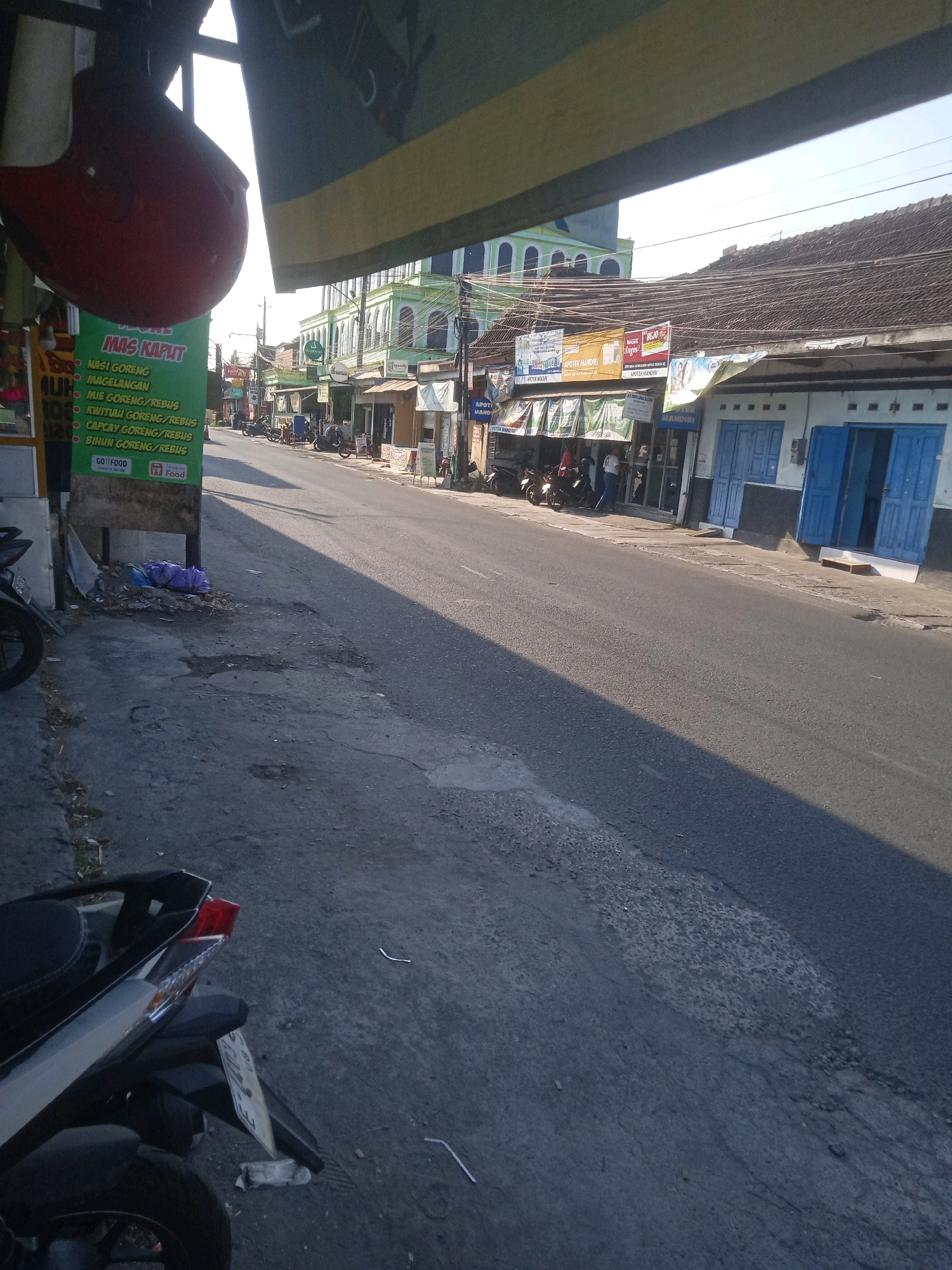 The image depicts a street scene in Trihanggo, Indonesia, likely during daylight hours given the bright sunlight and shadows. In the foreground, the rear of a parked motorcycle is visible, with its license plate partially obscured. To the left, a green sign with Indonesian text, possibly a menu or advertisement, hangs from a pole. Scattered debris and a purple plastic bag are present on the ground near the sign.
The street itself appears to be paved with asphalt and shows signs of wear and tear. On the right side of the street, buildings line the road. One building has blue double doors and a white facade. Further down the street, several shops with awnings are visible, and people can be seen in front of them. A green building with arched windows stands in the background. Overhead, power lines crisscross the sky, and a portion of a yellow and green awning extends into the frame from the top. The overall impression is of a typical urban street in a developing area.