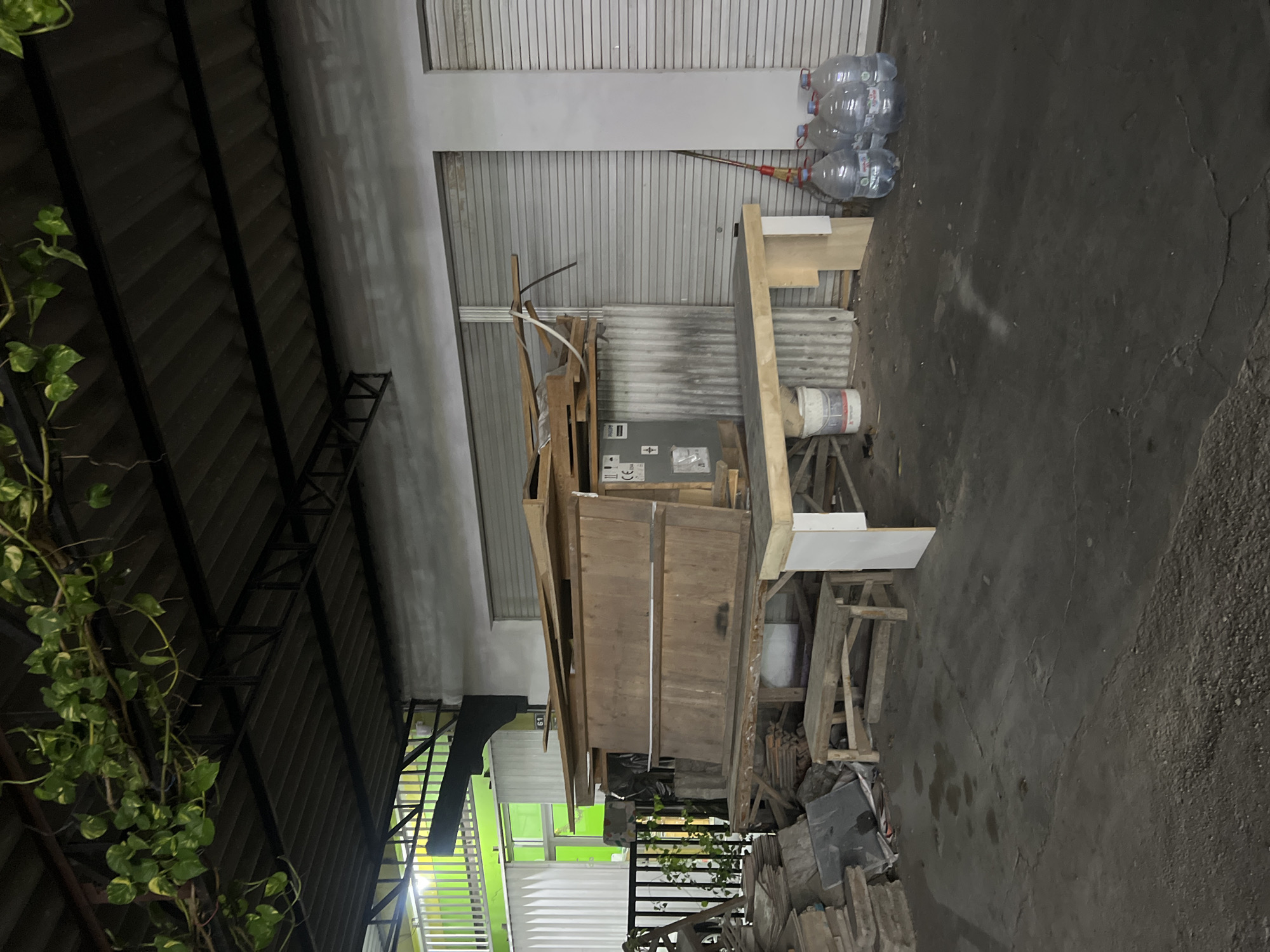 An outdoor storage area in Tlogoadi, Indonesia, is depicted in this medium shot. The scene is dominated by a large pile of lumber stacked on a wooden table supported by a workbench. To the left of the lumber, several pieces of slate or flat stone are piled on the ground, along with other debris. To the right of the lumber, five large plastic bottles of water are lined up on the ground. The area is covered by a corrugated metal roof supported by dark metal beams. Green ivy vines hang from the roof structure. In the background, there are two closed metal roll-up doors and a wall with a green illuminated window and vertical white siding. The ground is paved with concrete and appears wet in places, with scattered debris. A small wooden bench is situated beneath the lumber pile. The lighting suggests it is nighttime or dusk.