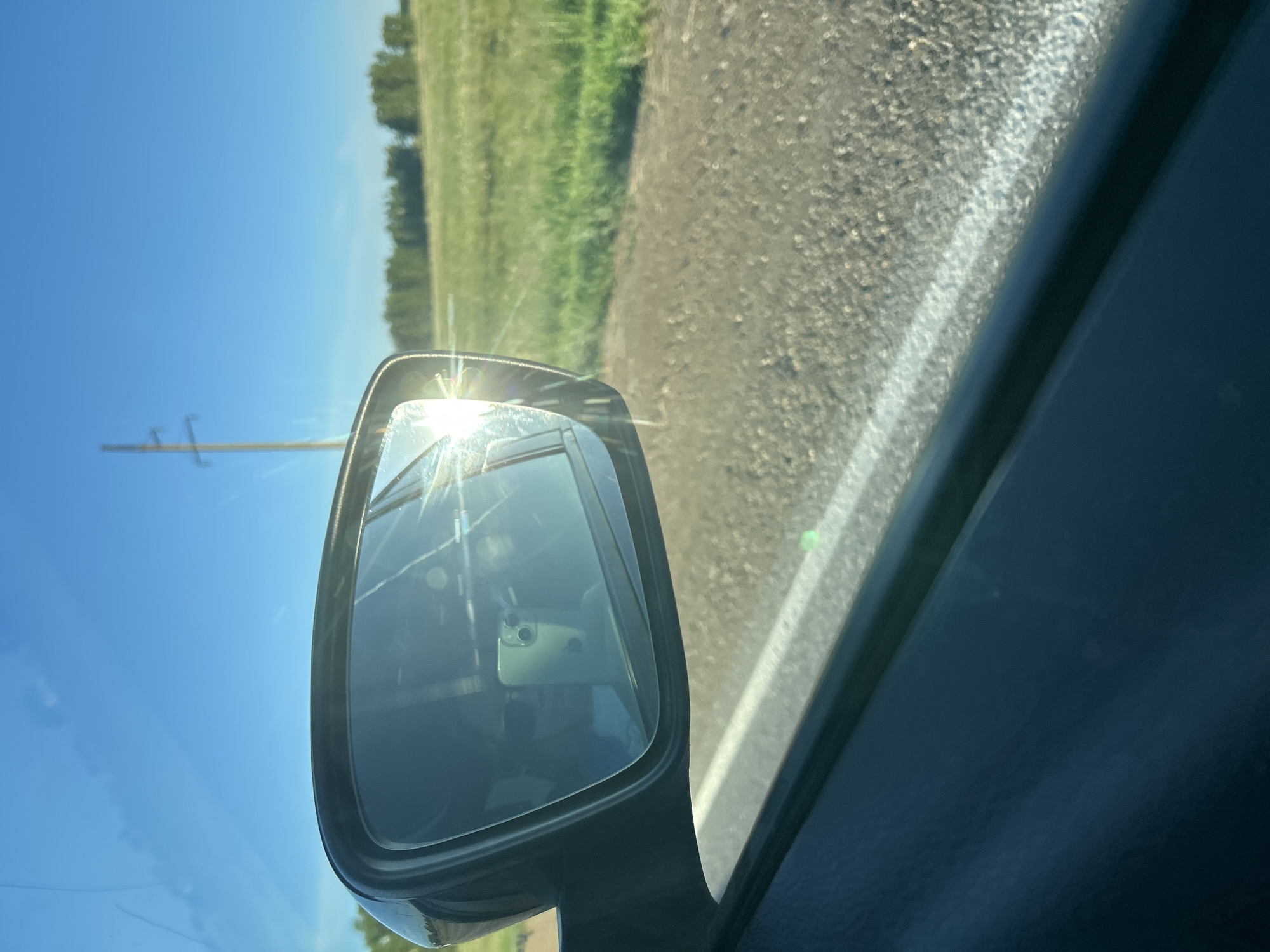 An outdoor, medium shot captures a side-view mirror of a vehicle parked on a dirt road, with a bright sun glare reflecting off the mirror. In the distance, a tree line borders a grassy field, and a utility pole stands against a clear blue sky. The interior of the car is visible in the mirror's reflection, showing a person wearing sunglasses and holding a cell phone. The scene is set in Ishmurzino, Russia.