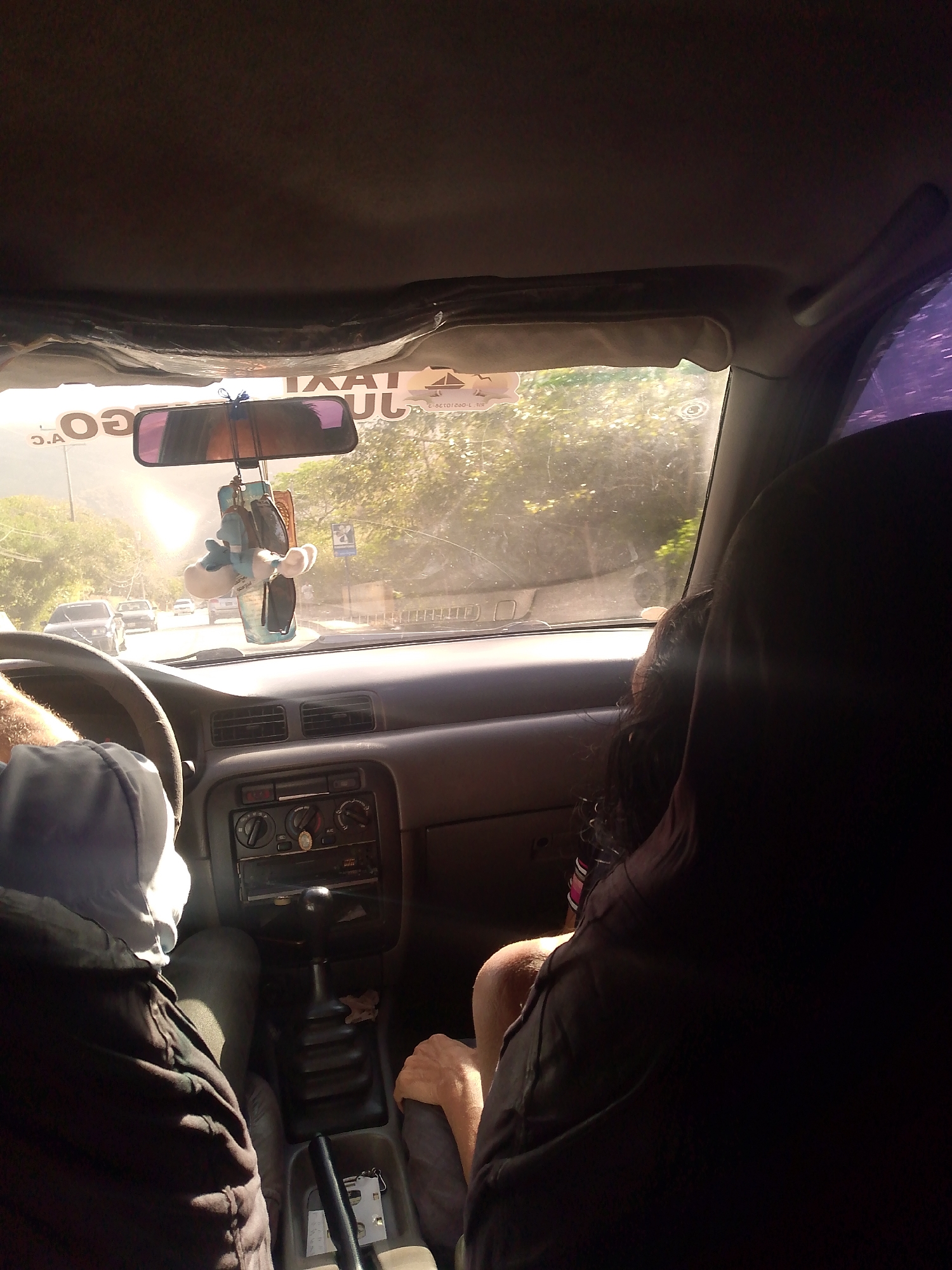 The scene is inside a taxi in La Asuncion, Venezuela. The driver is visible in the left foreground, wearing a hooded garment. A passenger sits in the front passenger seat. The dashboard of the car is visible, including the radio and gear stick. A rear-view mirror hangs from the windshield with a plush toy hanging from it. A "TAXI" sign is visible above the rear-view mirror. Outside, the landscape includes trees. Other cars are seen on the road in the distance.