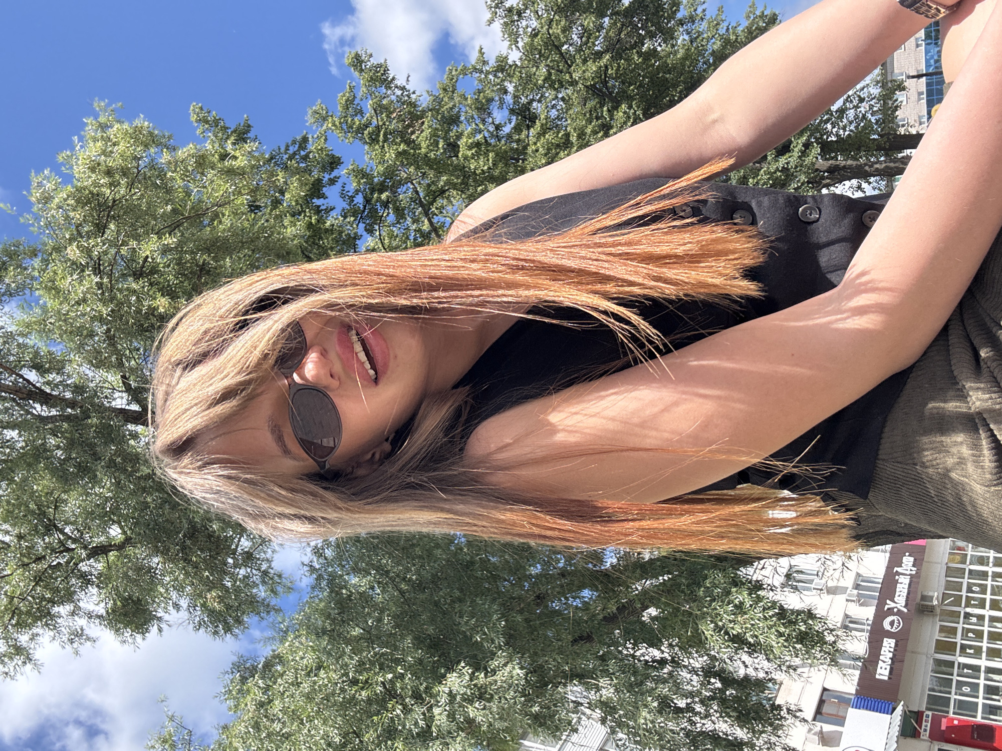 A young woman with long, blonde and orange-tinted hair and sunglasses smiles at the camera from a low angle, with a bright blue sky and fluffy white clouds in the background. She is wearing a black vest and a green corduroy skirt. Behind her, a large green tree is visible, as well as a building with a sign that reads "Пекарня" (Bakery) and "Хлебный Дом" (Bread House). The scene appears to be outdoors in an urban setting, likely in Chaikovsky, Russia, on a sunny day.