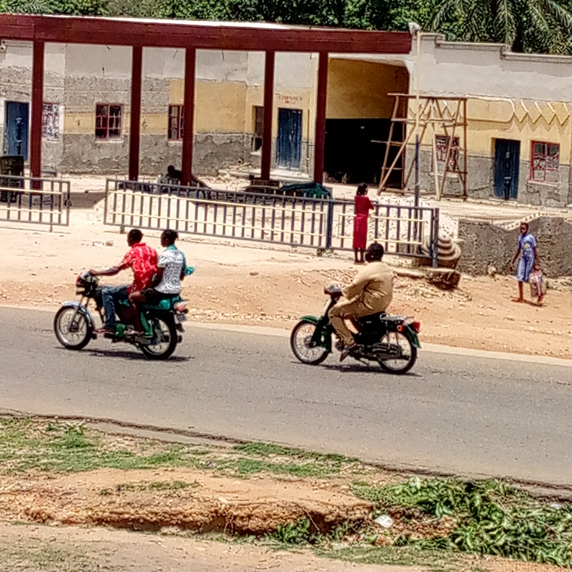 This outdoor image captures a bustling street scene in what is identified as Mararaba, Nigeria, under bright daylight.
In the foreground, an asphalt road runs horizontally across the bottom. Adjacent to the road, on both sides, are stretches of dry, reddish-brown earth and some scattered green vegetation.
The central part of the image features two motorcycles traveling on the road from left to right. On the left, a silver motorcycle carries two men; the front rider wears a red patterned shirt and blue jeans, while the passenger wears a white patterned shirt with a light blue piece of cloth or scarf over his shoulder. To their right, a single man, dressed in a light brown or tan outfit, rides a green motorcycle.
Behind the road and the motorcycles, a dirt area extends towards a large building in the background. A metal fence or barrier, painted in black and white or light and dark stripes, separates this dirt area from a foreground space that is out of frame.
The building in the background appears to be a multi-section structure with a weathered facade. It features multiple rectangular columns supporting a flat roof or overhang, all painted a dark reddish-brown. The walls behind these columns are a mix of gray concrete and faded yellow and white paint. There are several windows with dark frames and at least two blue doors visible. To the far right of the building, two wooden scaffolding structures or ladders are erected against the wall, suggesting ongoing construction or maintenance. Behind the building, dense green foliage, likely palm trees, is visible against the bright sky.
Several people are visible in the middle and background. To the left of the building's center, a woman in a long red dress stands near the metal fence, looking towards the right. Further to the right, in the dirt area, a person dressed in a blue patterned outfit walks away from the building, carrying a striped bag in their right hand.
The overall scene depicts a typical daily street environment with vehicular and pedestrian traffic, and a multi-purpose building, likely a commercial or public structure, suggesting an active community setting. No clearly readable text is visible on any signs or objects in the image.