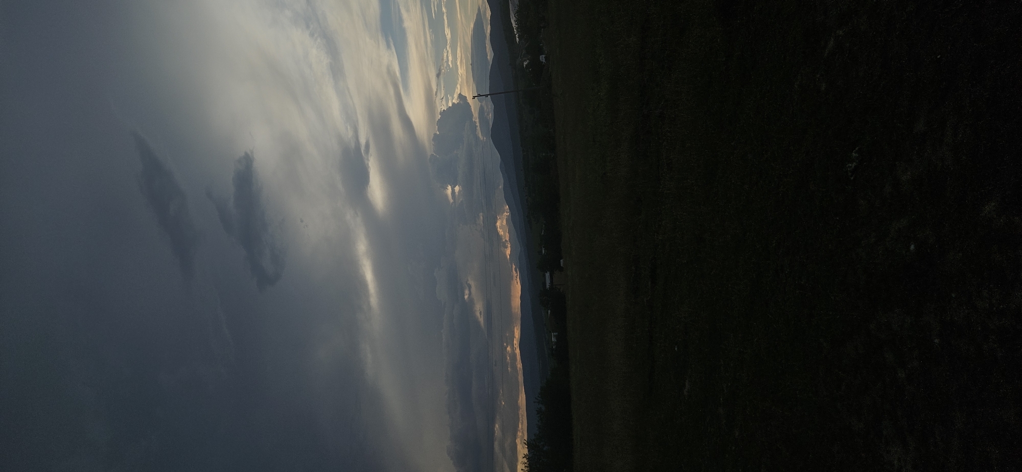 The image captures a dramatic landscape scene, likely at sunrise or sunset, in Momin Prohod, Bulgaria. The sky dominates the upper portion, featuring a striking contrast between dark, heavy storm clouds on the left and brighter, sun-kissed clouds towards the right. Golden and orange light from the obscured sun pierces through gaps in the cloud cover, illuminating their undersides and creating luminous streaks that highlight the layered texture of the clouds.
A range of mountains or hills stretches across the horizon, appearing silhouetted in deeper blues and grays against the brighter sky. Portions of these distant peaks catch the warm light, exhibiting soft orange and pink hues. A slender utility pole stands prominently against the mountains and sky on the right side, with faint power lines possibly visible.
The foreground and midground consist of a large, dark field or grassy area, cast mostly in shadow due to the low-angle lighting, which emphasizes the expansive sky and distant features. There are no people or animals visible, and no discernible activity is taking place beyond the natural weather phenomena. No text is visible in the media file. The overall impression is one of powerful natural beauty and changing weather conditions.