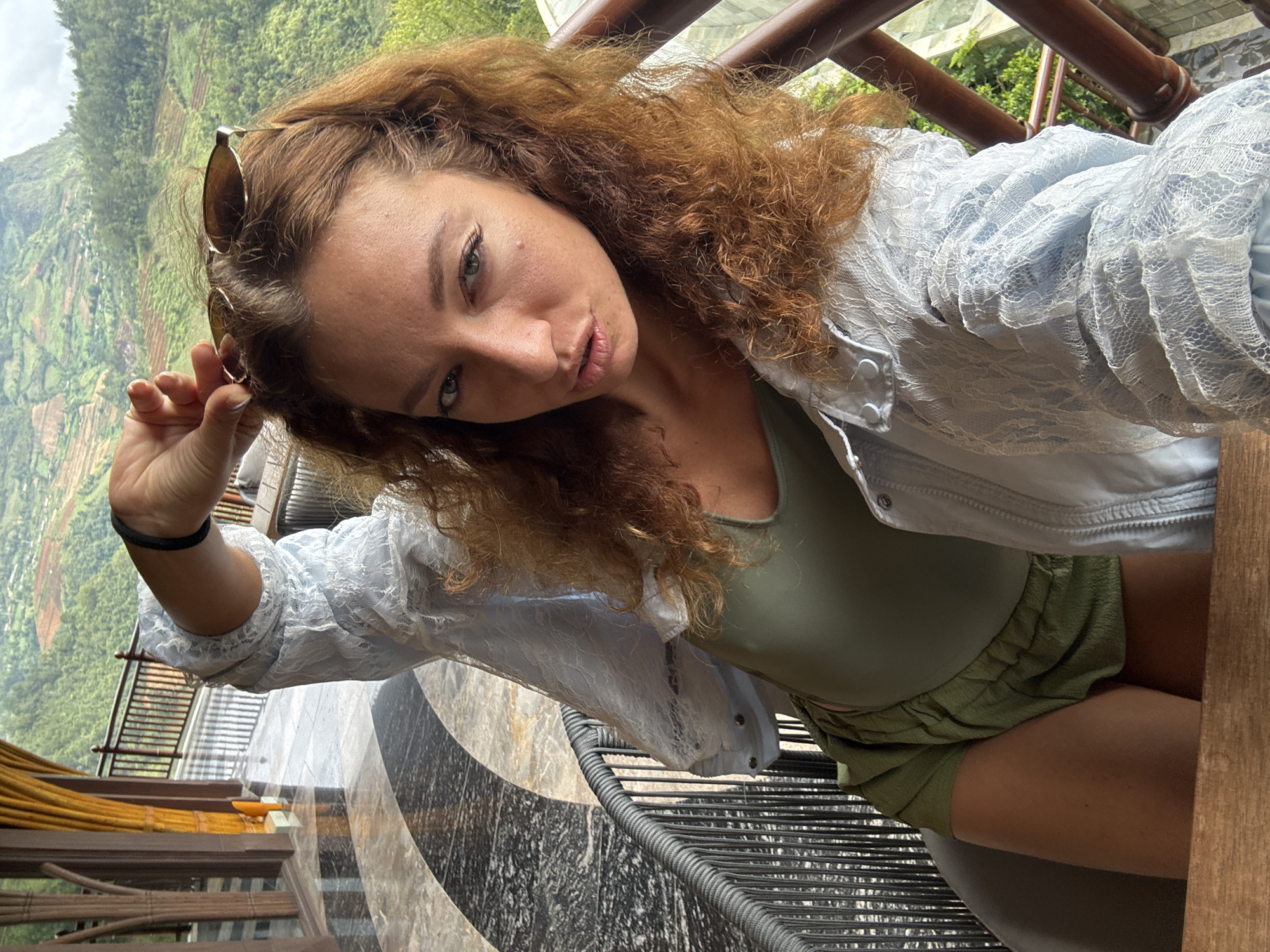 A selfie captures a young woman from a slightly high, tilted angle, gazing directly at the camera with a relaxed expression. Her long, reddish-brown curly hair frames her face, and sunglasses are pushed up onto her head. She wears a light blue, sheer lace jacket over an olive green sleeveless top and olive green shorts. Her left hand is visible, holding the arm of her sunglasses.
The woman is positioned on what appears to be a balcony or terrace, with intricate black and white patterned floor tiles beneath her. Various wooden and metal railings, possibly part of a balustrade or furniture, are visible in the foreground and around her. To her right, a dark, woven outdoor chair or furniture piece is partially visible.
In the background, a breathtaking mountainous landscape unfolds, characterized by extensive green terraced fields cascading down the slopes. This scenery is consistent with the mountainous regions of Vietnam, specifically Púng Luông. The lighting suggests it is daytime, with bright ambient light illuminating the scene. There is no visible text in the image.