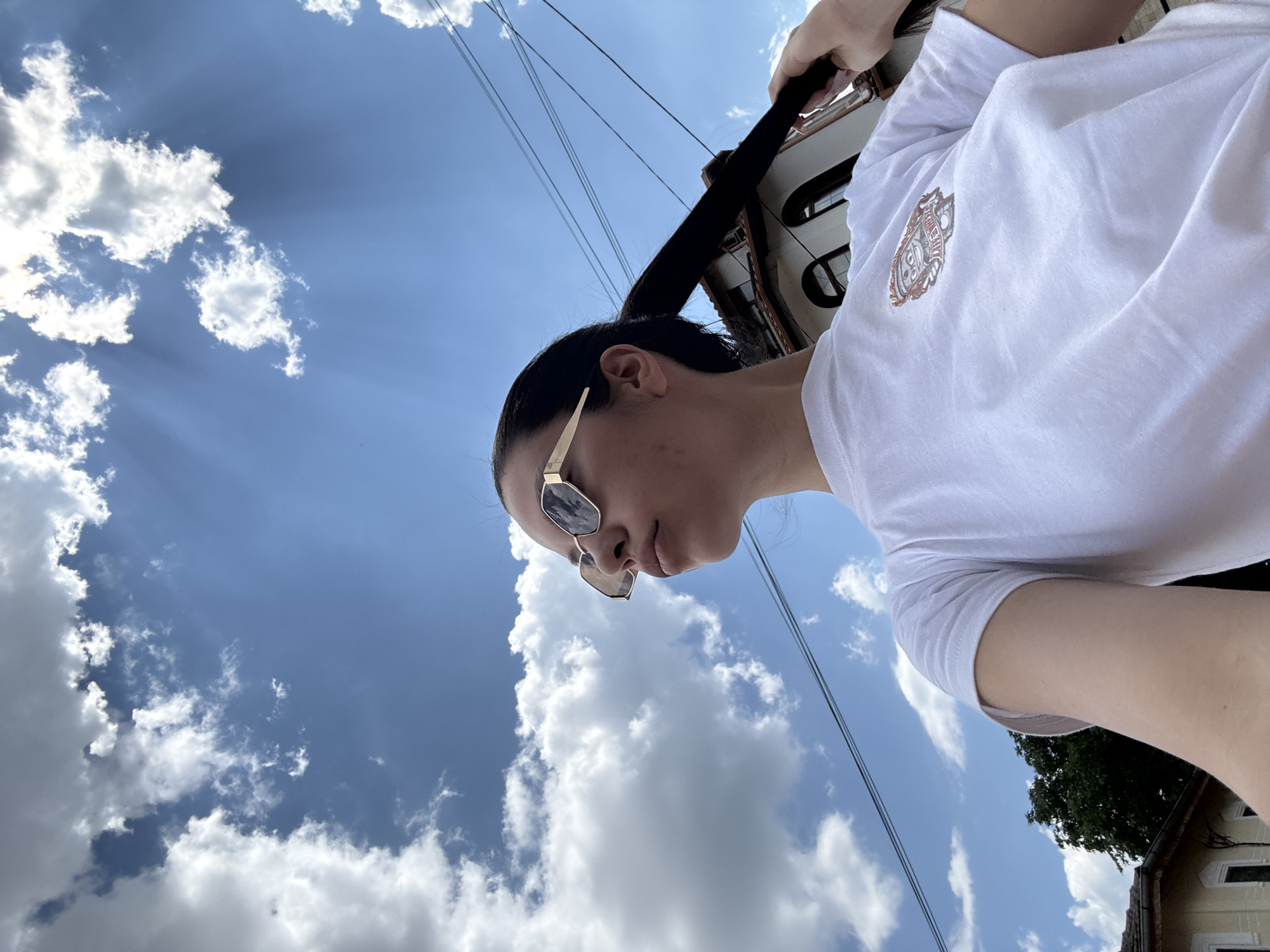 In Chavdar, Bulgaria, a young woman with dark hair pulled back into a ponytail, wearing sunglasses and a white t-shirt, poses for a selfie outdoors. She is looking up and to her left, with her right hand holding her ponytail. The background features a bright blue sky with scattered white clouds, the upper portion of a building with arched windows, and overhead power lines. A tree is visible on the left side of the frame.