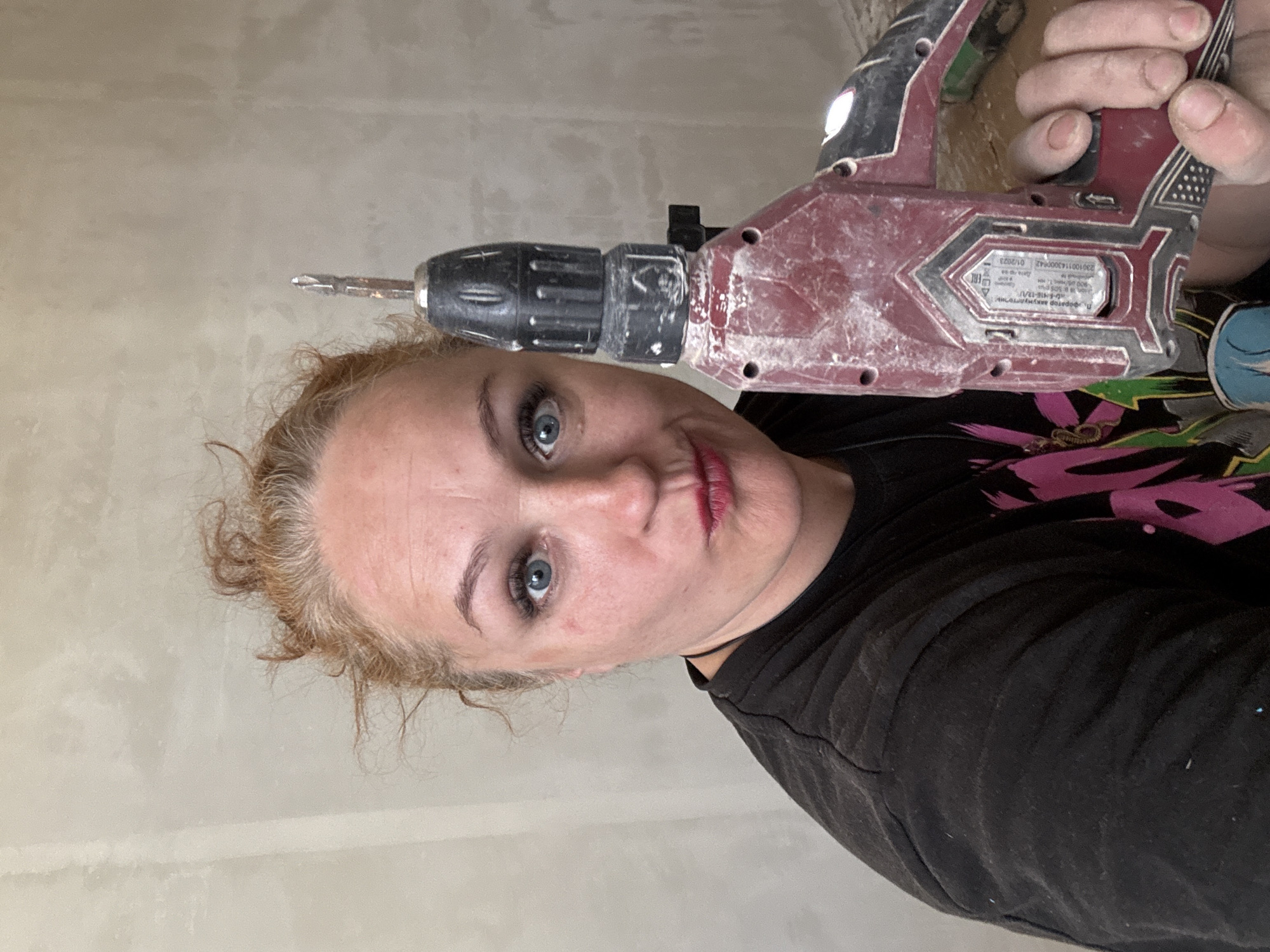 A fair-skinned person with blonde hair and blue eyes is holding a drill. The drill is red and black. The background is a light beige wall. The person is wearing a black shirt. The location is possibly Chaikovsky, Russia, based on the drill's markings and manufacturer.
