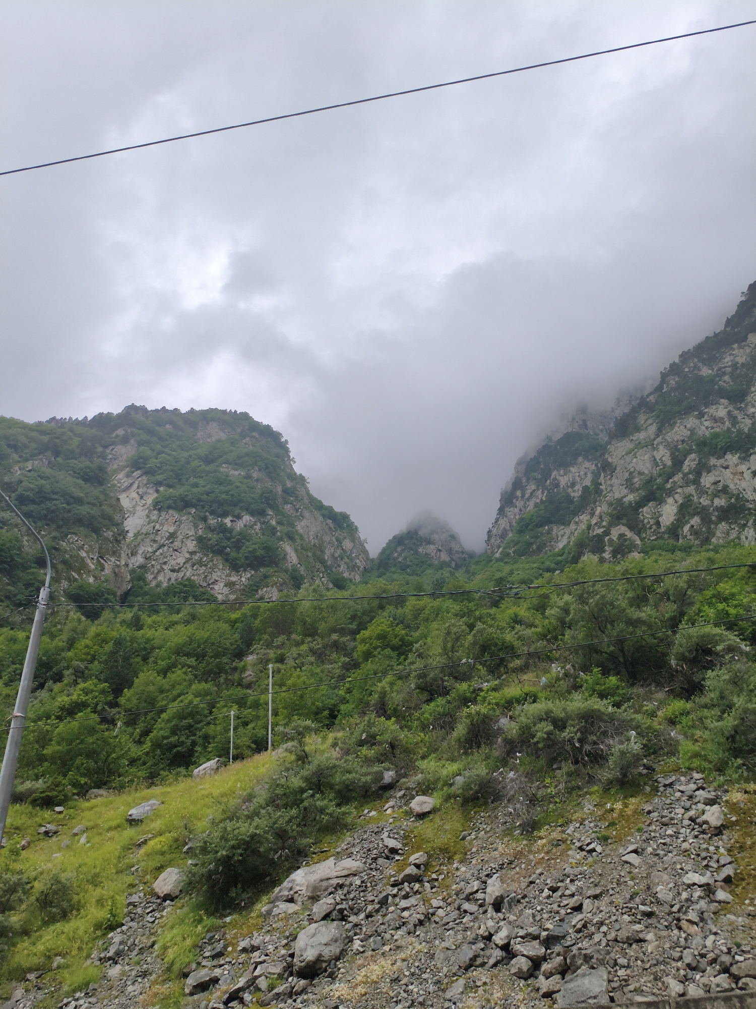 The image depicts a mountainous landscape near Verkhnii Lars, Russia. A prominent power line stretches across the cloudy sky. The slopes are covered with dense green vegetation, rocks, and scree. In the foreground, a rocky, grassy slope is visible, with a light-colored utility pole on the left. In the background, steep, rocky mountains rise, partially obscured by mist and clouds, suggesting a potentially wet or overcast weather condition. The overall scene conveys a rugged, natural environment.