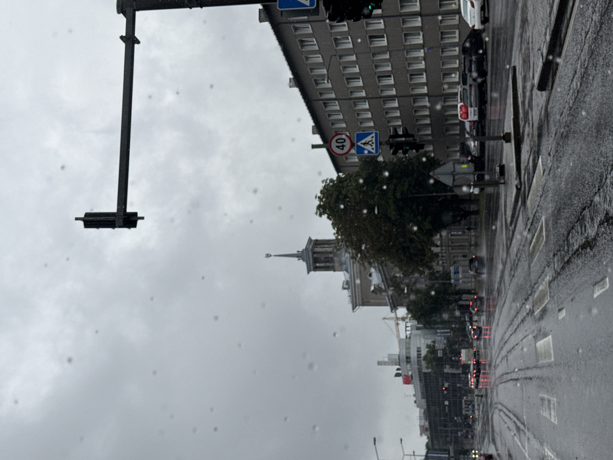 The image depicts a city street in Tallinn, Estonia, on a rainy day. The street is wet and reflects the lights of vehicles. Several cars are visible, driving on the wet asphalt. There are tram tracks on the road. Buildings line the street, including a multi-story building with many windows and a building with a distinctive spire. Traffic lights and road signs are present, including a speed limit sign indicating 40 and a pedestrian crossing sign. The sky is overcast and gray, with raindrops visible on the lens or in the foreground. The overall scene conveys a gloomy, wet atmosphere typical of rainy weather in an urban environment.