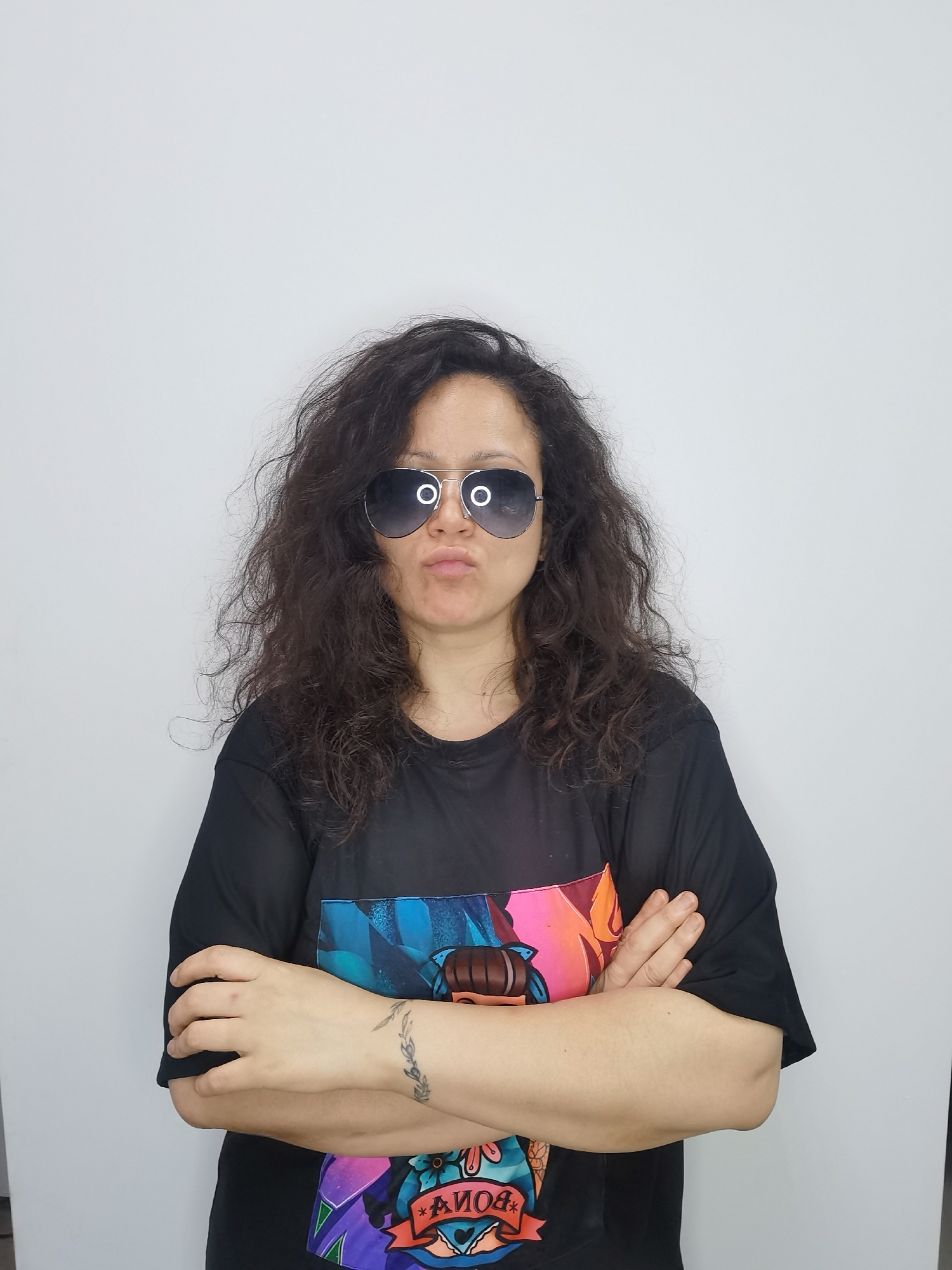 A woman with long, curly dark hair is shown from the chest up, wearing aviator sunglasses and a black t-shirt with a colorful graphic print. She has her arms crossed and her lips are pursed. She has a tattoo on her left wrist. The background is a plain white wall. The location is not identifiable within the city of Chaikovsky, Russia.