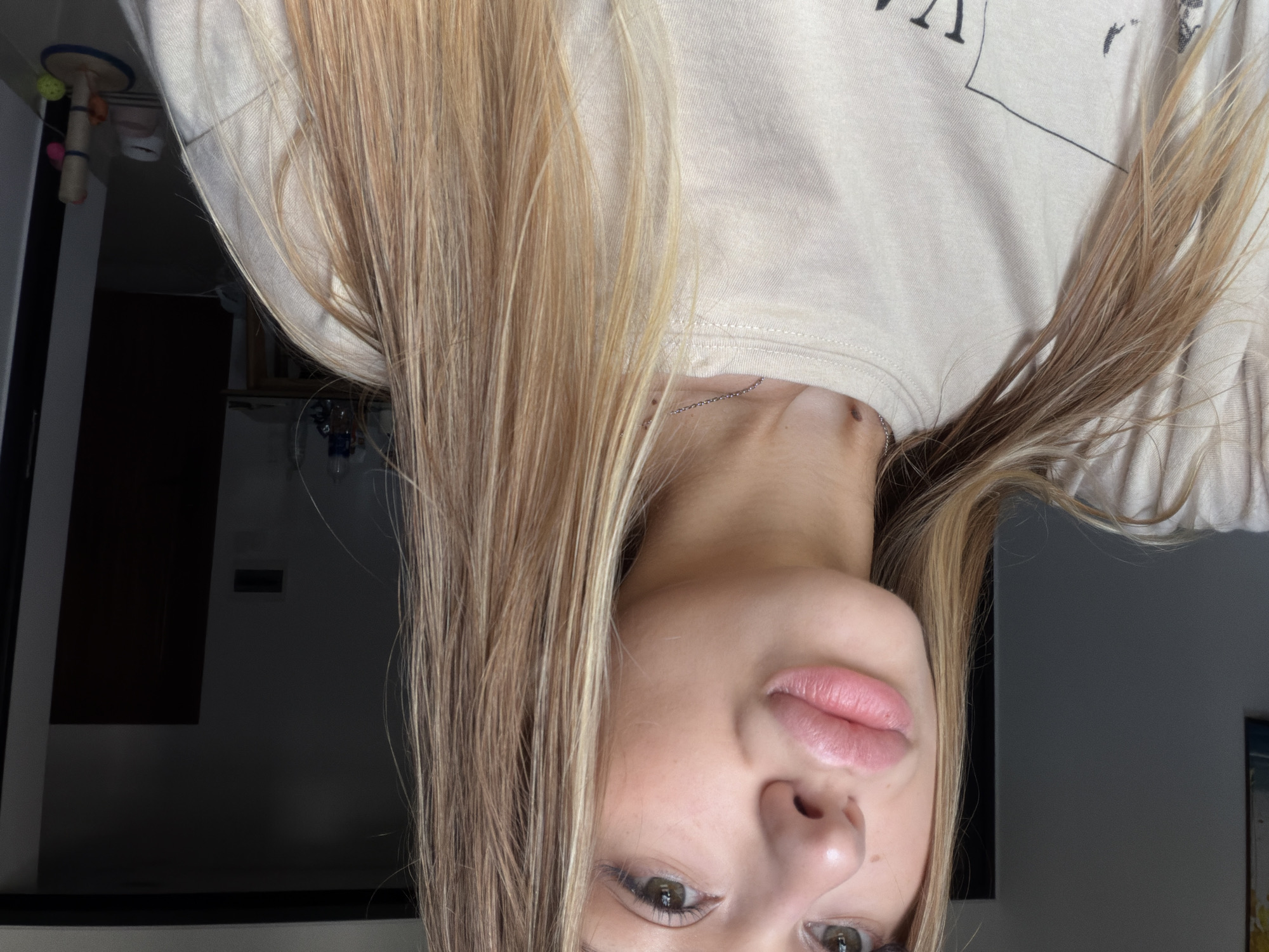 A close-up, head-and-shoulders shot of a young woman with blonde hair and green eyes is presented. She is looking directly at the camera with her lips pursed. She is wearing a light-colored t-shirt with a graphic design on it. The background is a dimly lit room with furniture visible. There is no indication that this scene is located in Đà Nẵng, Vietnam.