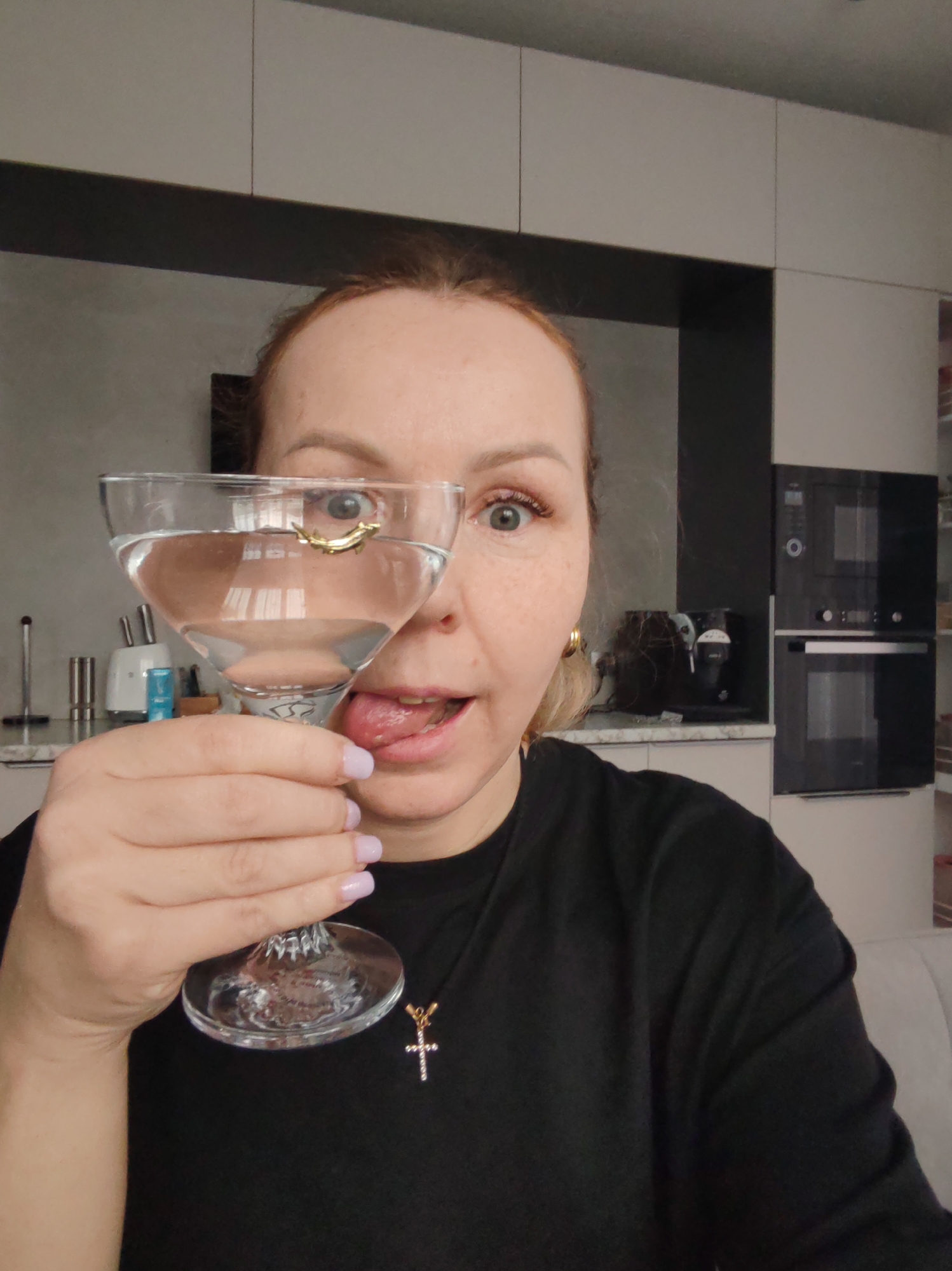 A woman is holding a martini glass filled with water, in which a gold-colored object, possibly a ring, is submerged. She is sticking her tongue out and making a playful face towards the camera. She is wearing a black shirt and a cross necklace. The background shows a modern kitchen with gray and white cabinets, built-in ovens, and various kitchen appliances. Based on metadata, the image was taken in Chaikovsky, Russia.