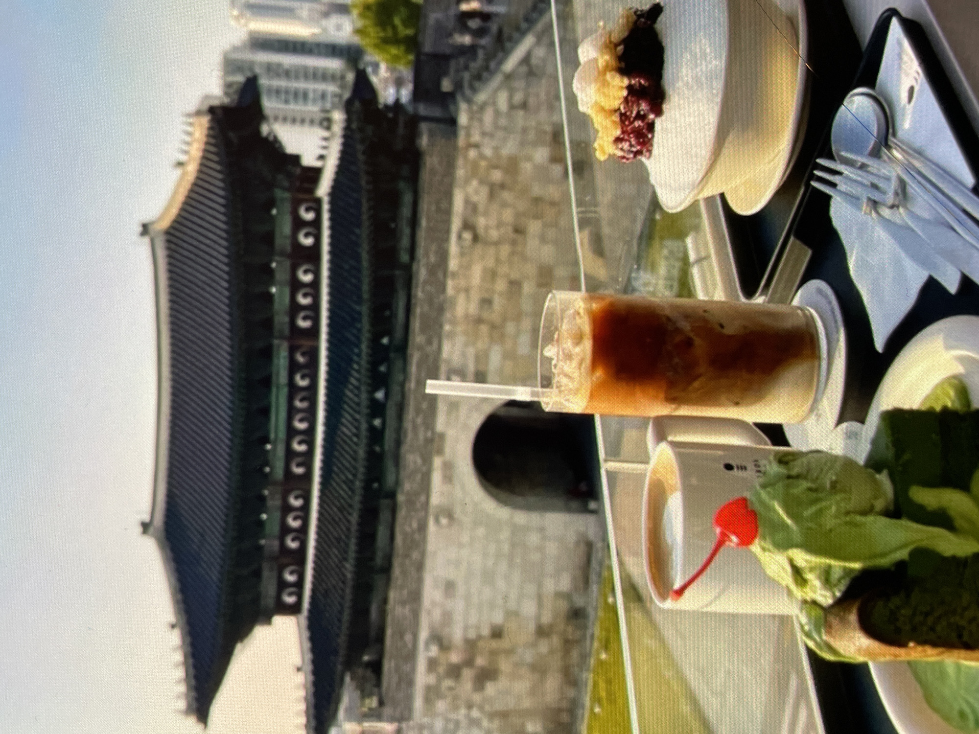 The image shows a table with food and drinks, including a dessert in a white bowl with red and yellow toppings, an iced coffee in a glass, a coffee cup with a red cherry on a spoon, and what appears to be a green dessert. There is also a spoon and fork on a napkin on a tray. In the background is a traditional Asian building with dark gray roof tiles and a light gray wall, along with modern buildings in the distance. The location is not in Harmanli, Bulgaria. The building is likely located in Asia.