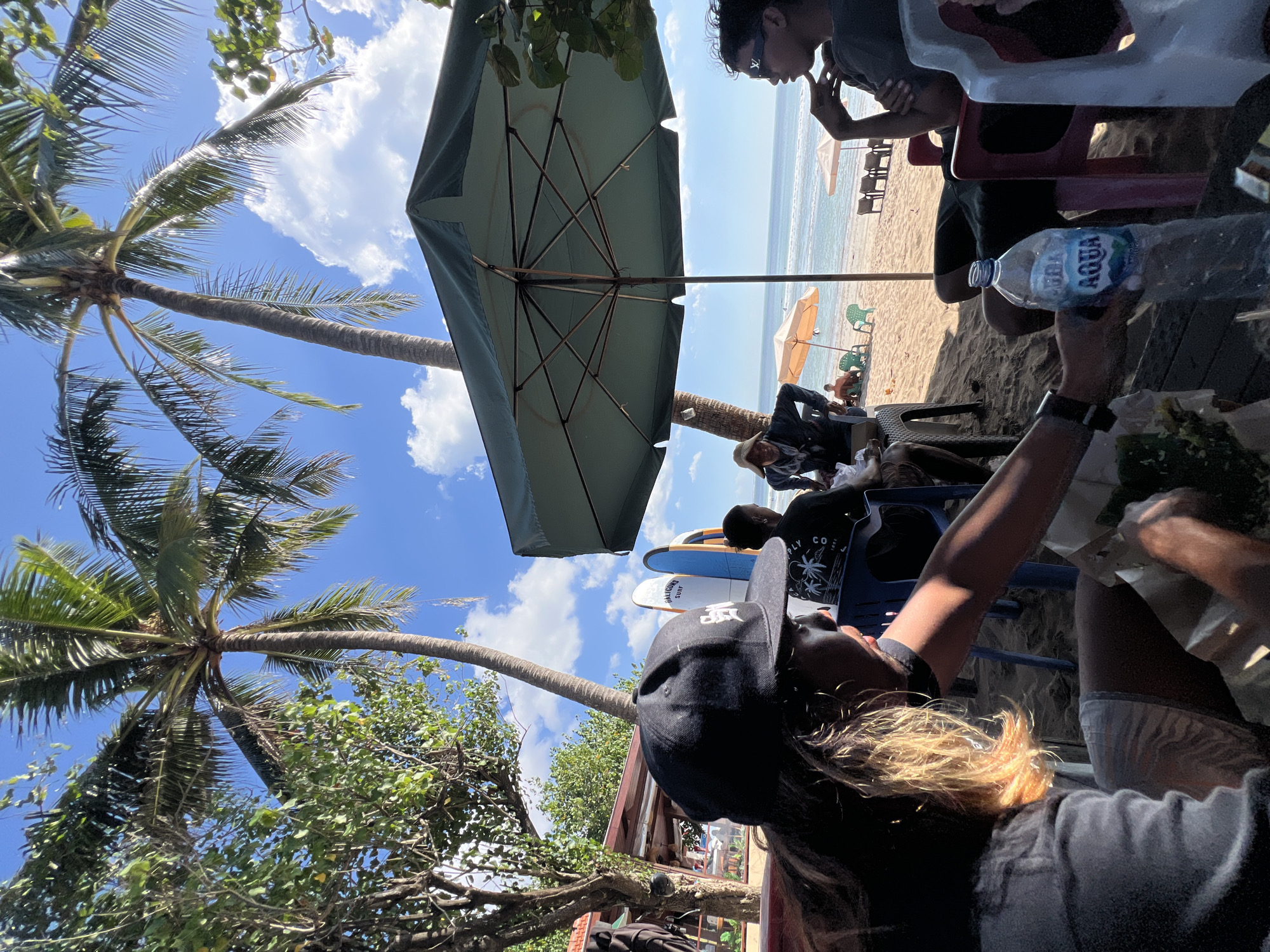 In Kuta, Indonesia, a group of people are gathered on a sandy beach, likely at a beachfront cafe, under a large green umbrella. The sun is shining brightly, casting shadows on the sand. Two palm trees tower over the scene, their fronds reaching towards a bright blue sky with scattered white clouds. In the foreground, a person wearing a black baseball cap and light-colored shorts is partially visible, holding a paper container of food. Next to them, a plastic bottle of Aqua water is placed on a table. To the right, another person with sunglasses is seated, looking towards the ocean. In the background, several surfboards are propped upright. Further down the beach, beach chairs and umbrellas are set up, and a few individuals are relaxing. The ocean stretches out to the horizon, with gentle waves lapping at the shore. The overall atmosphere appears relaxed and casual, typical of a beachside location in Kuta.