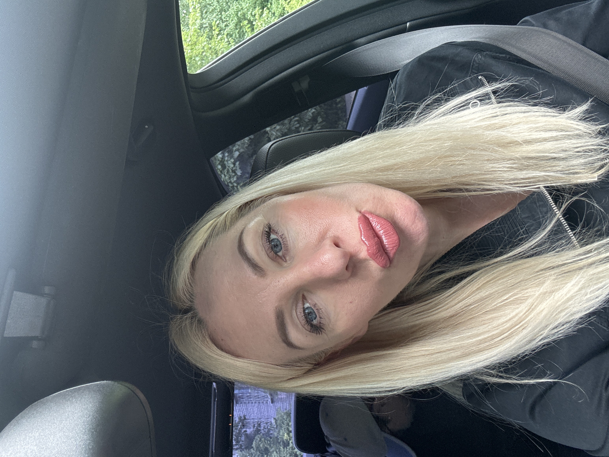The image shows a close-up of a woman with blonde hair and blue eyes, tilted to the side. She is wearing a black jacket and has noticeable makeup, including pink lipstick. The background suggests she is inside a car, with glimpses of greenery and trees through the window. The lighting and the overall composition suggest it might be daytime. There is no visible text in the image. The scene is set in Saint Petersburg, Russia.
