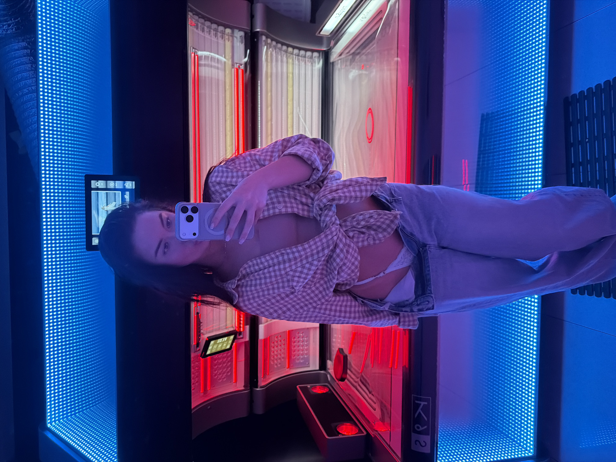 A young adult female holds a cell phone in a tanning booth in Moscow, Russia. The booth is illuminated with red lights, and the walls of the surrounding room have blue LED panels. The female is wearing jeans and an unbuttoned, long-sleeved, checkered shirt. The image appears to be a selfie taken with the cell phone.