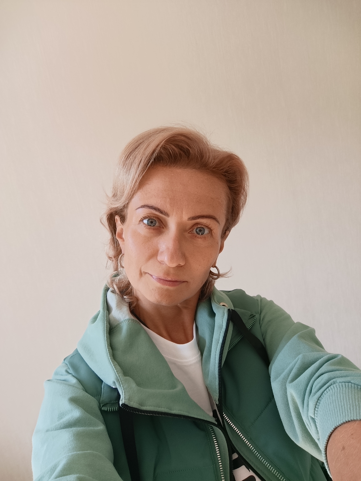 The image is a selfie of a woman with short, blonde, wavy hair, light blue eyes, and fair skin. She is wearing a light green zip-up jacket over a white t-shirt. She is looking directly at the camera with a slight smile. The background is a plain, light-colored wall. The image appears to be taken indoors, likely in a residential setting. There is no information in the image to suggest it is located in Moscow, Russia.