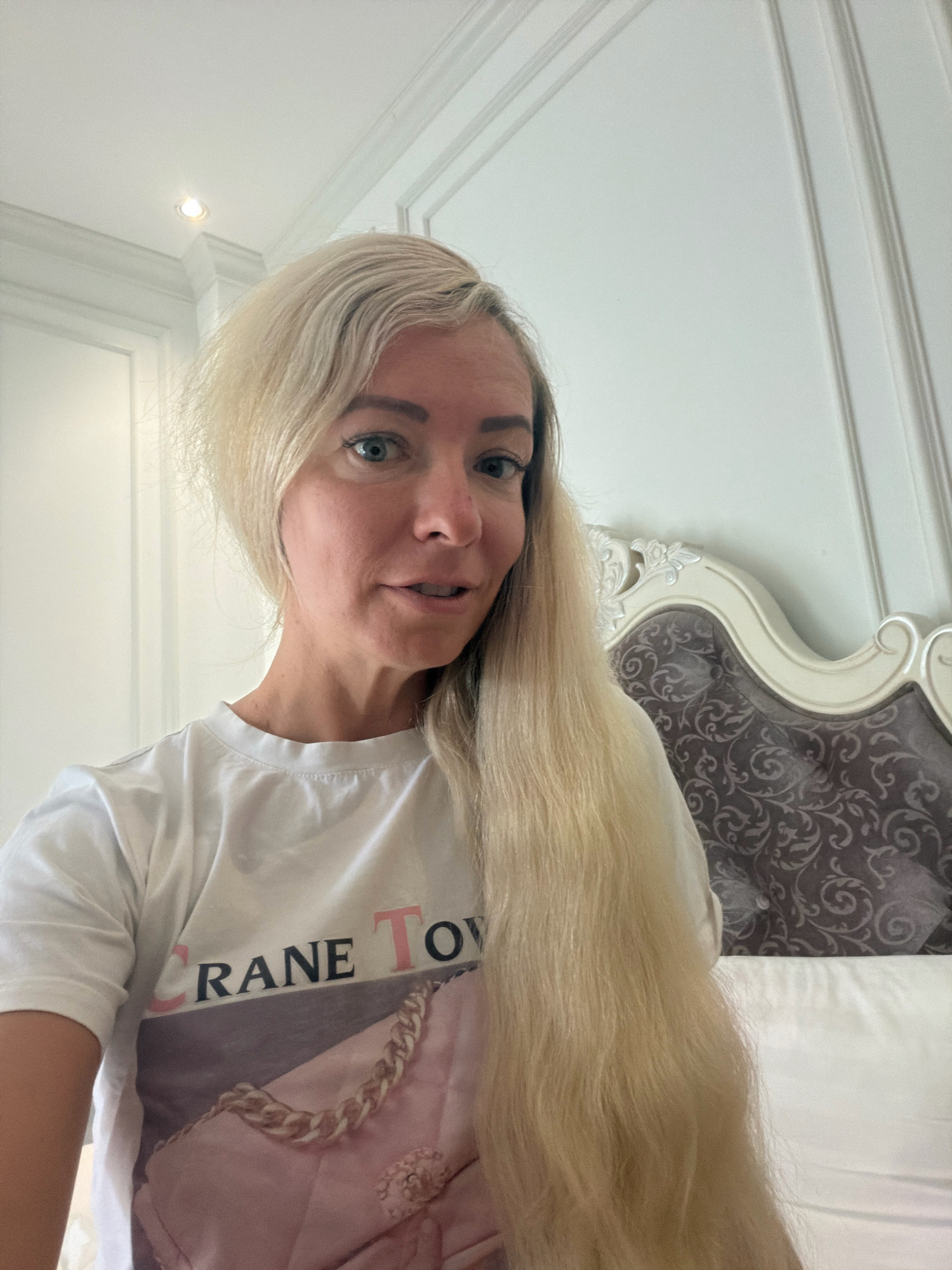 A fair-skinned woman with blonde hair, wearing a white t-shirt with a pink handbag graphic and "CRANE TOWN" text, is shown from the chest up. She has blue eyes and is looking towards the camera with her mouth slightly open. Her hair is long and falls over her right shoulder. The background consists of a white wall with decorative molding and a portion of a headboard with ornate carvings and patterned upholstery. The lighting suggests an indoor setting, possibly a bedroom. There is no visual information in the image that allows for identification of the city of Xuân Thành, Vietnam.