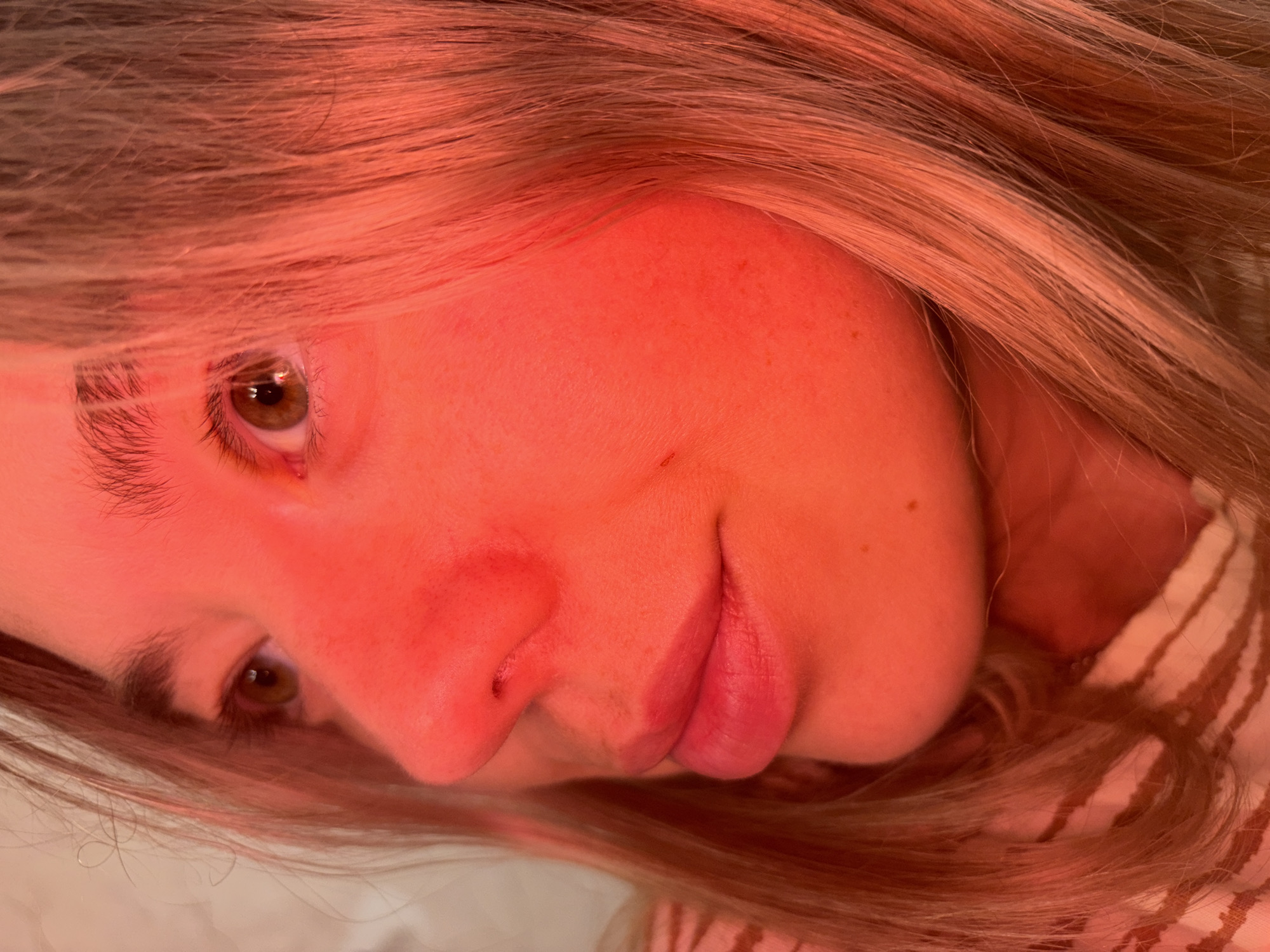 The image displays a close-up, three-quarter profile portrait of an individual, likely female, facing towards the upper right of the frame. The scene is bathed predominantly in a strong, artificial red light, which saturates the subject's skin, hair, and visible clothing in tones of red, orange, and deep pink.
The subject's left eye (viewer's left) is clearly visible, with a light brown or hazel iris and a dilated pupil, reflecting a small, bright light source. Eyelashes are discernible, and a dark eyebrow is present above the eye. The right side of the subject's face is partially obscured by strands of light-colored hair, which appear reddish-orange due to the lighting. The nose is prominent, and the left nostril is visible. The lips are full, slightly parted, and appear to have a soft pink or reddish hue, likely from lipstick, also influenced by the red illumination. The skin texture is visible, with minor imperfections such as small moles or freckles on the cheek and chin areas.
The hair is long and appears to be blonde or light brown in natural light but is heavily tinted red and orange here. It flows around the face and shoulders, with individual strands distinguishable. A segment of patterned clothing, possibly striped in off-white and brown tones, is visible on the subject's lower left side. The background is out-of-focus and appears to be a light, indistinct surface, rendered in shades of muted pink and orange by the prevailing red light, suggesting an indoor environment. No objects or specific environmental markers are discernible to indicate a precise location or context beyond the subject herself.