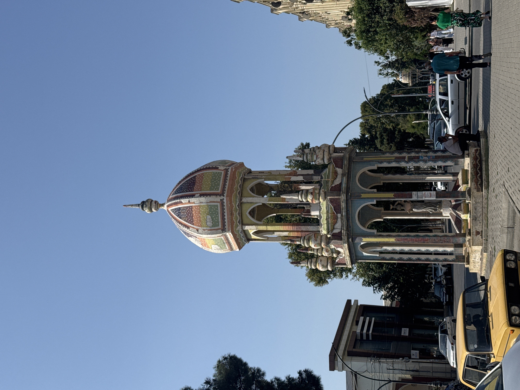 In Khachmaz, Azerbaijan, a mosaic-covered, multi-tiered fountain stands in the foreground under a clear blue sky. The fountain features arched openings, decorative spires, and a colorful, domed roof adorned with intricate patterns. To the left, a modern building with dark horizontal accents is visible, along with parked cars, including a distinctive yellow sedan. The street is paved with cobblestones, and trees line the background. Several people are walking and sitting, some near the fountain, and others on a sidewalk in the distance. The scene appears to be a public space within the city.