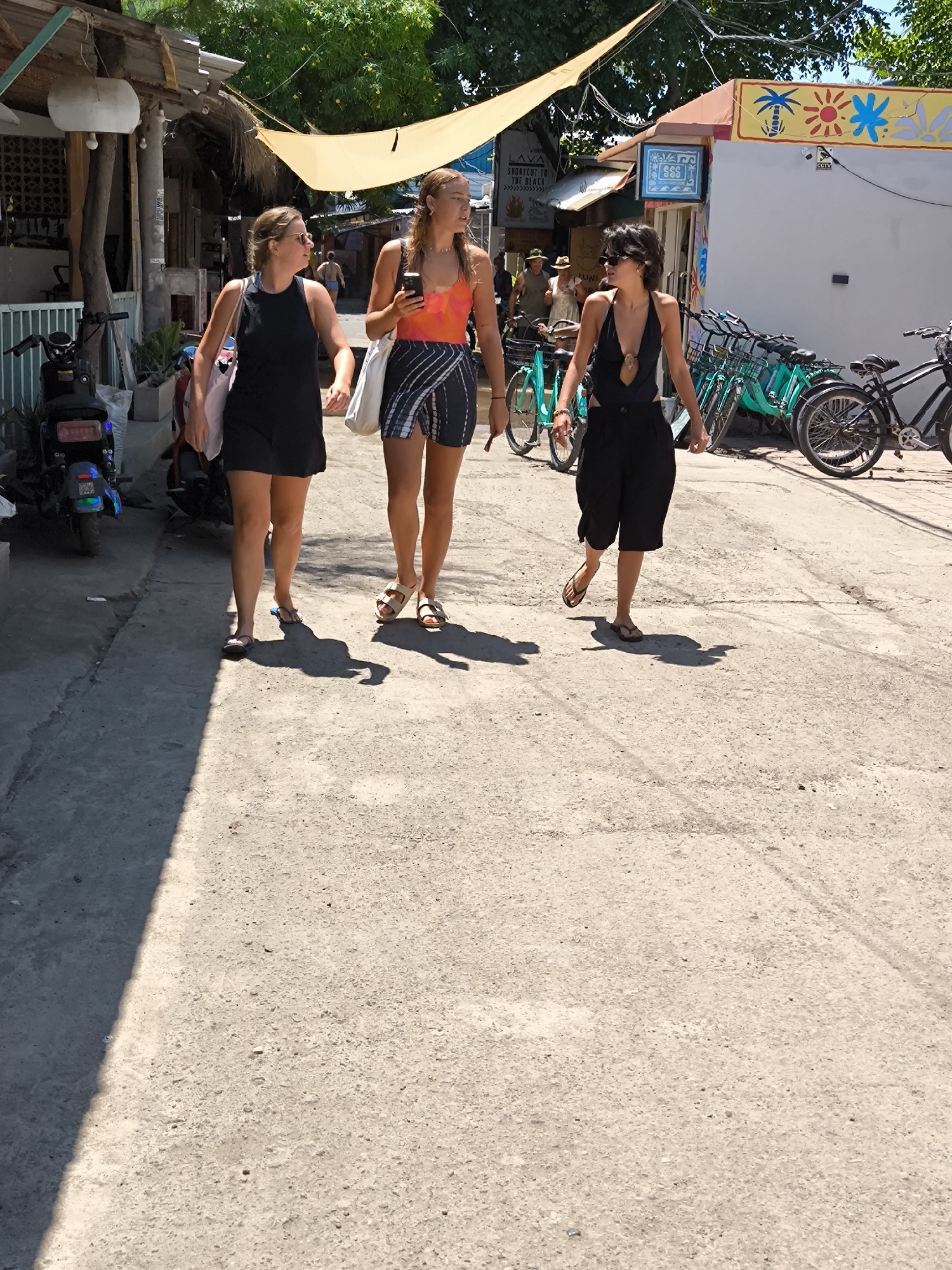 Three women are walking down a street in what appears to be a tropical location. The woman on the left is wearing a black dress, sunglasses, and carrying a tote bag. The woman in the middle is wearing a colorful top and a sarong, and is looking at her phone. The woman on the right is wearing a black halter top and shorts, also with sunglasses. The street is lined with shops and bicycles. There is a scooter parked on the left side of the street. A light colored awning is strung above the street. The setting appears to be a street in Gili Trawangan, Indonesia.