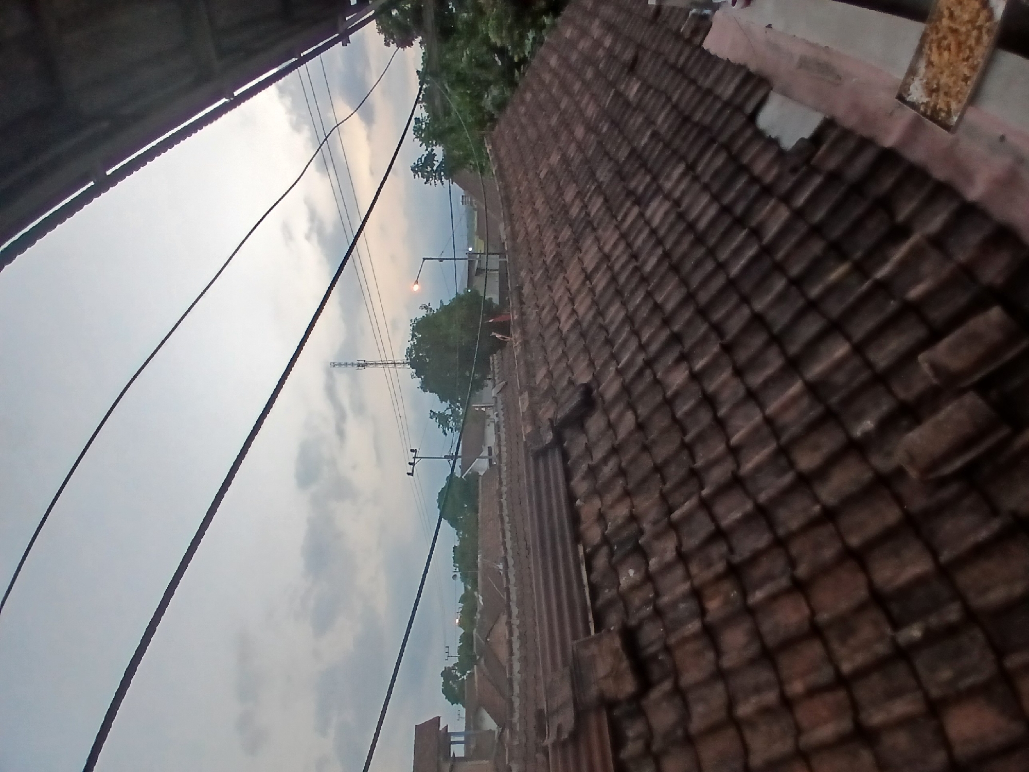 The image captures a view of rooftops and overhead wires in the city of Tenggarang, Indonesia, under a cloudy sky, possibly during twilight. The foreground is dominated by a close-up of weathered, reddish-brown terracotta roof tiles, some of which appear damaged or dislodged. Beyond the immediate rooftops, the scene opens up to reveal a cluster of other houses with similar tiled roofs, interspersed with lush green trees. In the middle ground, a tall telecommunications tower stands prominently against the sky. A streetlamp is visible, its light on, suggesting it is dusk or early evening. Electrical wires are strung across the sky, adding a utilitarian layer to the urban landscape. In the far right foreground, a rectangular tray filled with what appears to be fried rice or a similar food item is placed on a ledge. The overall impression is of a residential area in an Indonesian city, characterized by traditional roofing materials and the presence of modern infrastructure like power lines and communication towers.