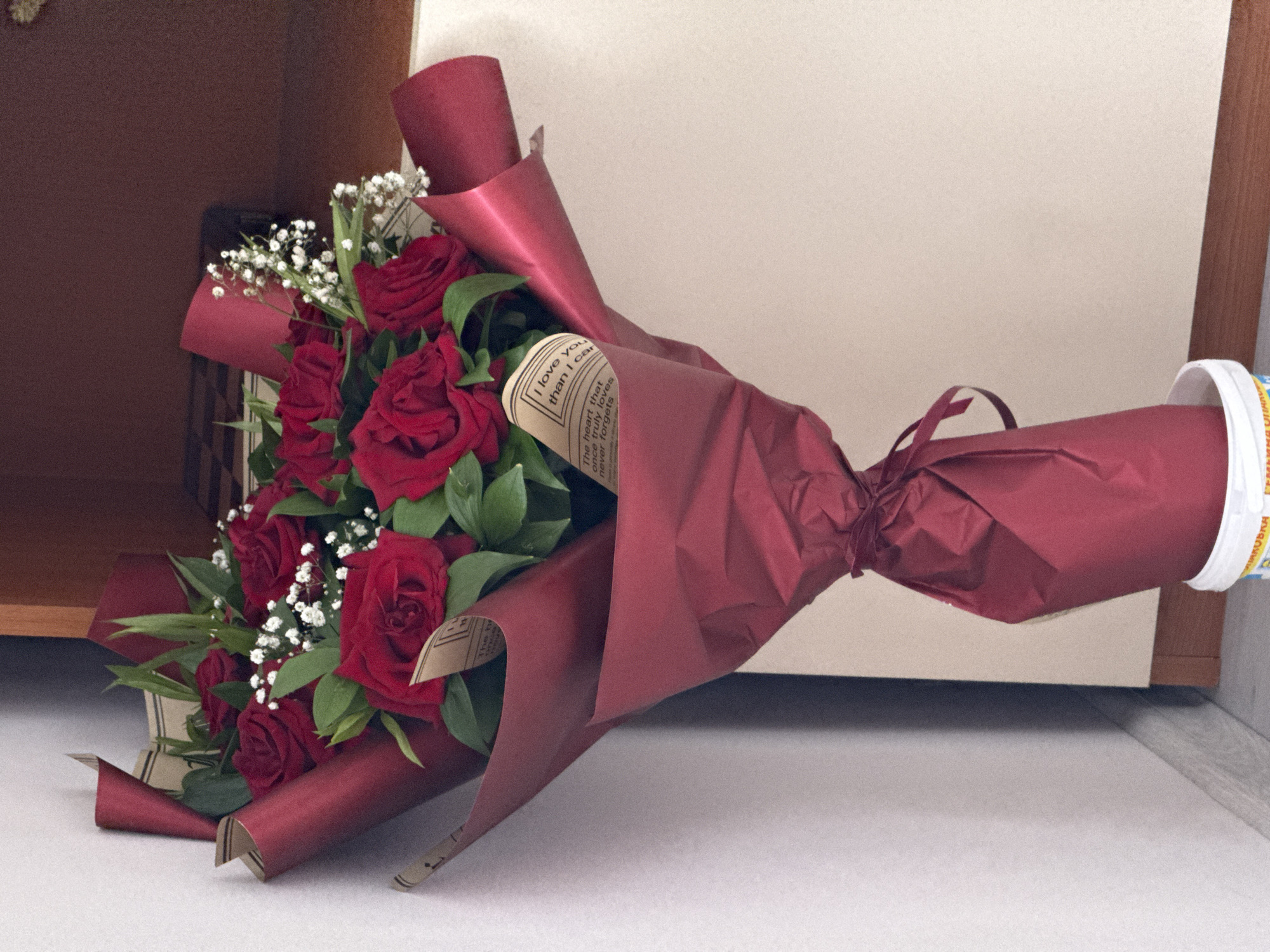 A vibrant bouquet of red roses, adorned with white baby's breath and green foliage, is the central subject of the image. The roses are a deep, rich red, and the bouquet is intricately wrapped in a luxurious maroon-colored paper, tied with a matching ribbon around the stems.
Part of the wrapping includes a lighter, beige-toned paper that features visible English text. Phrases such as "I love you," "than I can," "The heart that," "one truly loves," and "never forgets" are discernable, suggesting a romantic or affectionate message.
At the very end of the bouquet's stem, a cylindrical object with a white rim and a yellow section is partially visible, featuring Cyrillic text that appears to read "...ЕМЫШКА...". This could be part of a branded wrapper or container.
The bouquet is resting on a light-colored, possibly white or light grey, flat surface, suggesting an indoor setting. The background consists of a plain, light-toned wall or panel, with a darker brown, possibly wooden, surface visible on the left side of the frame. There are no people present, and no specific activity is taking place; the bouquet is stationary, presented as a gift or decoration. The overall impression is one of love, celebration, or appreciation. No specific time of day or weather conditions can be determined from the indoor setting, nor are there explicit visual cues relating to the Varna, Bulgaria location context.