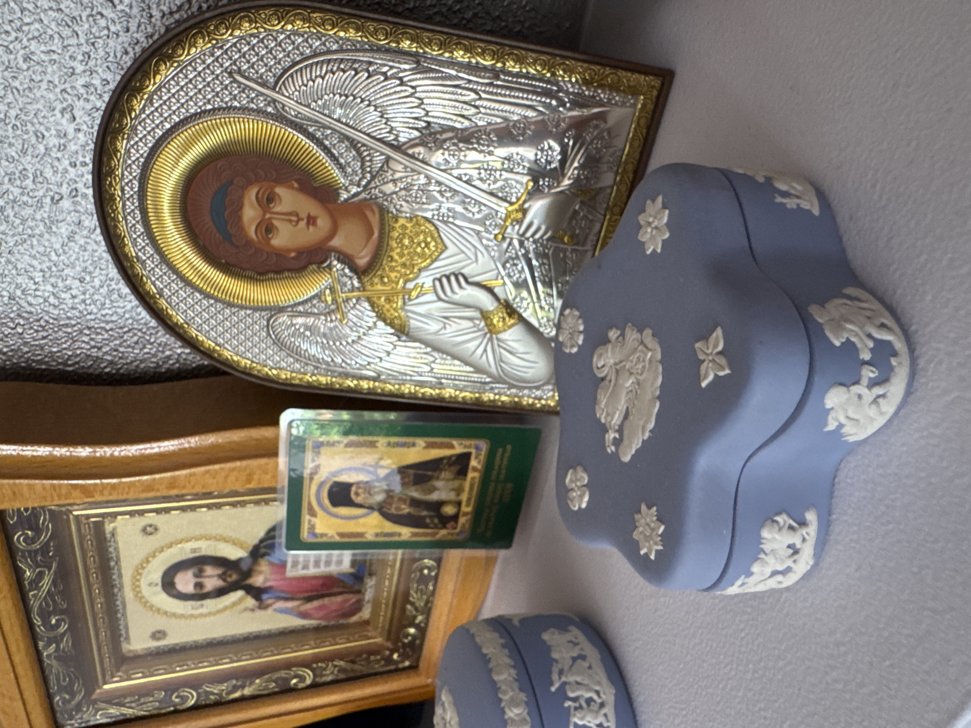 A still life photograph captures an arrangement of religious icons and decorative objects on a white surface, likely a shelf or table. In the background, two framed icons are visible. The icon on the left, in a wooden frame, depicts a figure with a halo and blessings. To its right, a smaller, rectangular icon with a green border shows a robed man with a black hat.
Dominating the center-right of the frame is a large, arched icon of an angel, possibly Michael, rendered in silver and gold. The angel has wings, a halo, and holds a staff and sword.
In the foreground, two ornate, powder-blue ceramic boxes with white bas-relief figures are arranged. The box on the left is round, and the one on the right has a distinctive quatrefoil shape. These boxes feature classical motifs, including cherubic figures and floral designs. The overall impression is one of devotional items and decorative artistry. The location of Yeysk, Russia, is not discernible from the image itself.