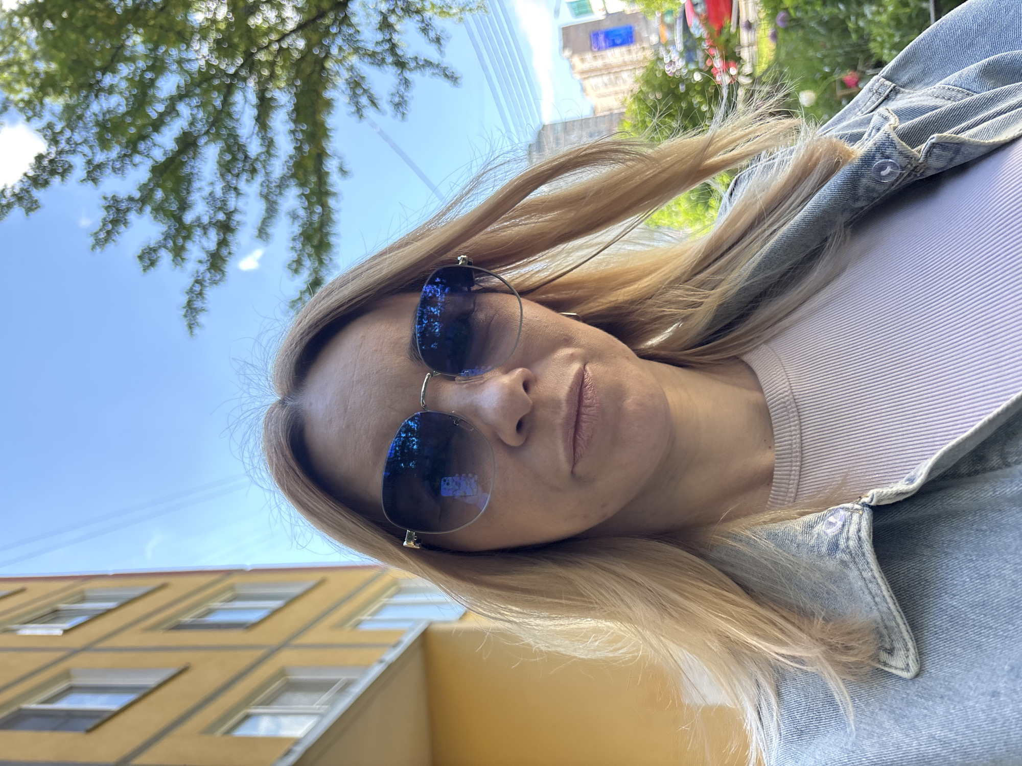The image displays a woman taking a selfie, with the photograph oriented at an approximately 90-degree angle. The subject is a woman with long, blonde hair, some strands of which appear windswept. She is wearing large, reflective sunglasses that obscure her eyes, a light-colored (possibly light pink or lavender) ribbed top, and a light blue denim jacket. Her mouth is slightly parted, and her overall expression appears neutral.
The background is bright and sunny, indicating daytime. Above her head and to the left, a clear blue sky is visible, partially covered by blurred green foliage, suggesting trees. In the upper right, blurred buildings with some colorful features are present. To the lower left, a portion of a distinct yellow building with multiple rows of rectangular windows is visible. The reflective surface of her sunglasses shows blurred distortions of the surrounding environment, likely buildings and the sky. There is no visible text in the image. The scene suggests an outdoor setting in what appears to be an urban or suburban environment.
