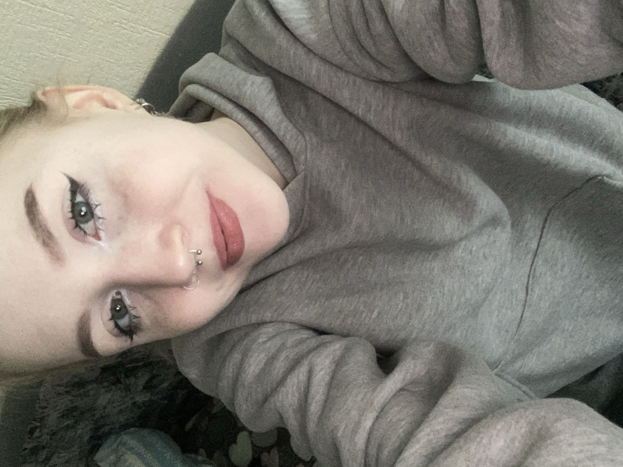 A young woman lies on a surface covered with a mottled gray and black pattern, possibly a blanket or a cushion. She is wearing a gray hooded sweatshirt. The woman's face is the focal point, angled towards the camera. Her makeup includes winged eyeliner and dark lipstick. She has piercings on her nose and ear. The background is blurred and indistinct, appearing to be a beige wall. There is no evidence to support that the location is Izhevsk, Russia.