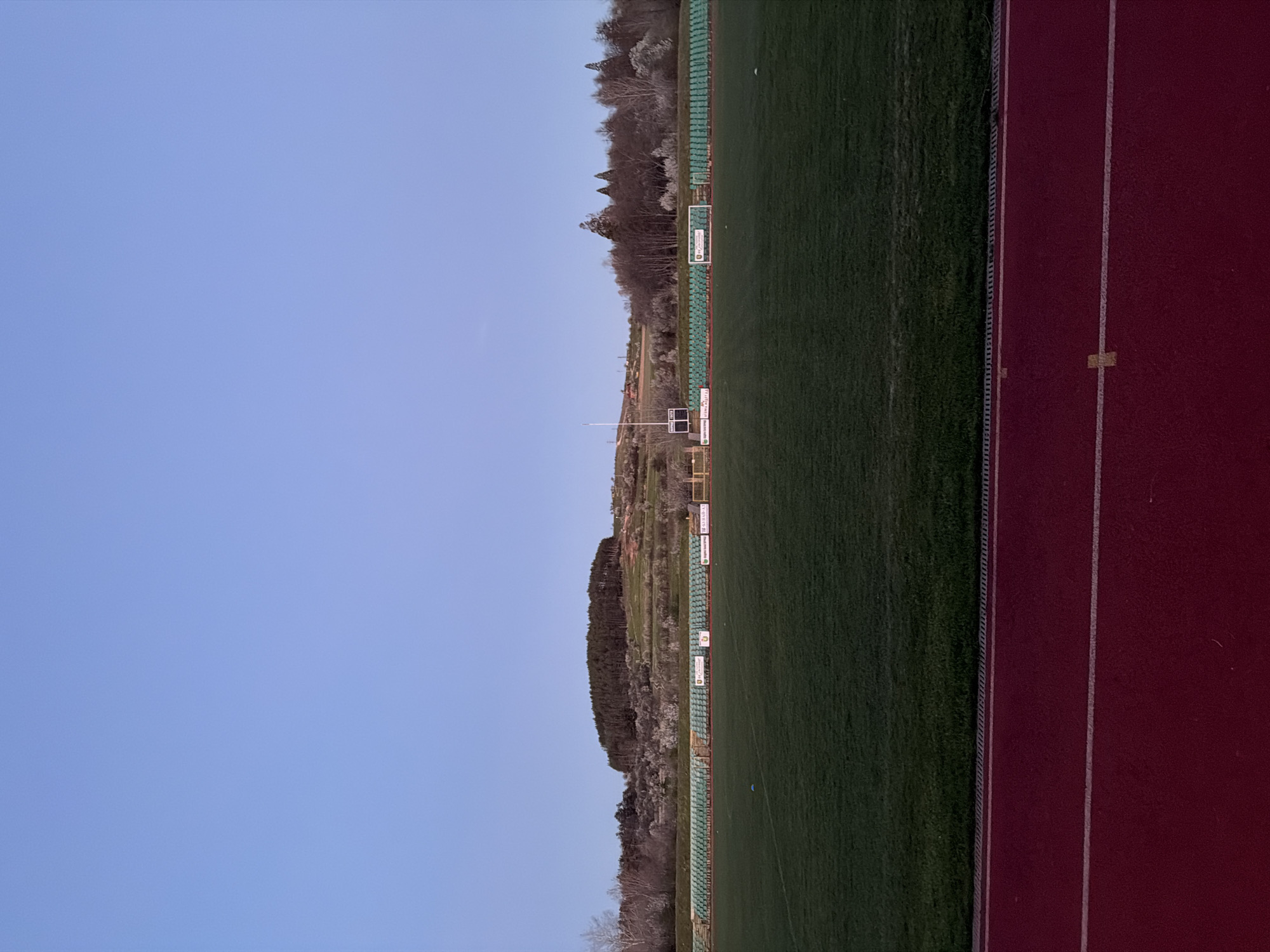 The image depicts a stadium in Panagyurishte, Bulgaria. On the right is a red running track with white lane markings. Adjacent to the track is a green grass field. Behind the field are rows of green stadium seating, with advertisements mounted on the back. A scoreboard is present near the center of the image. A wooded area forms the background. The sky is blue and clear. The image is oriented vertically.