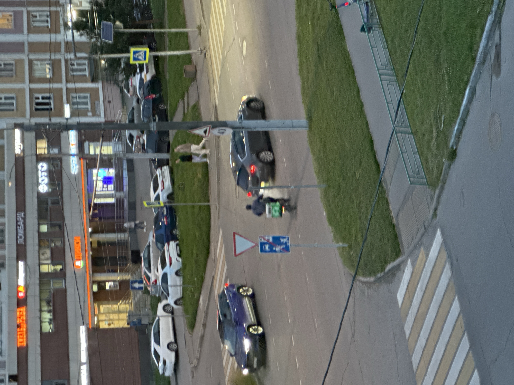 An aerial view captures a street scene in Podolsk, Russia. A blue SUV is turning at an intersection, its headlights illuminated. To its right, a dark sedan with its brake lights on is also in the intersection, and a person on a scooter with a green box is proceeding through it. Across the street, a pedestrian crossing is visible. In the background, buildings line the street, with businesses displaying illuminated signage. One sign reads "FOTO," and others are in Cyrillic script. The sky appears overcast, and the lighting suggests it is late afternoon or early evening. Green grass borders the road and sidewalk, and a metal fence is present.