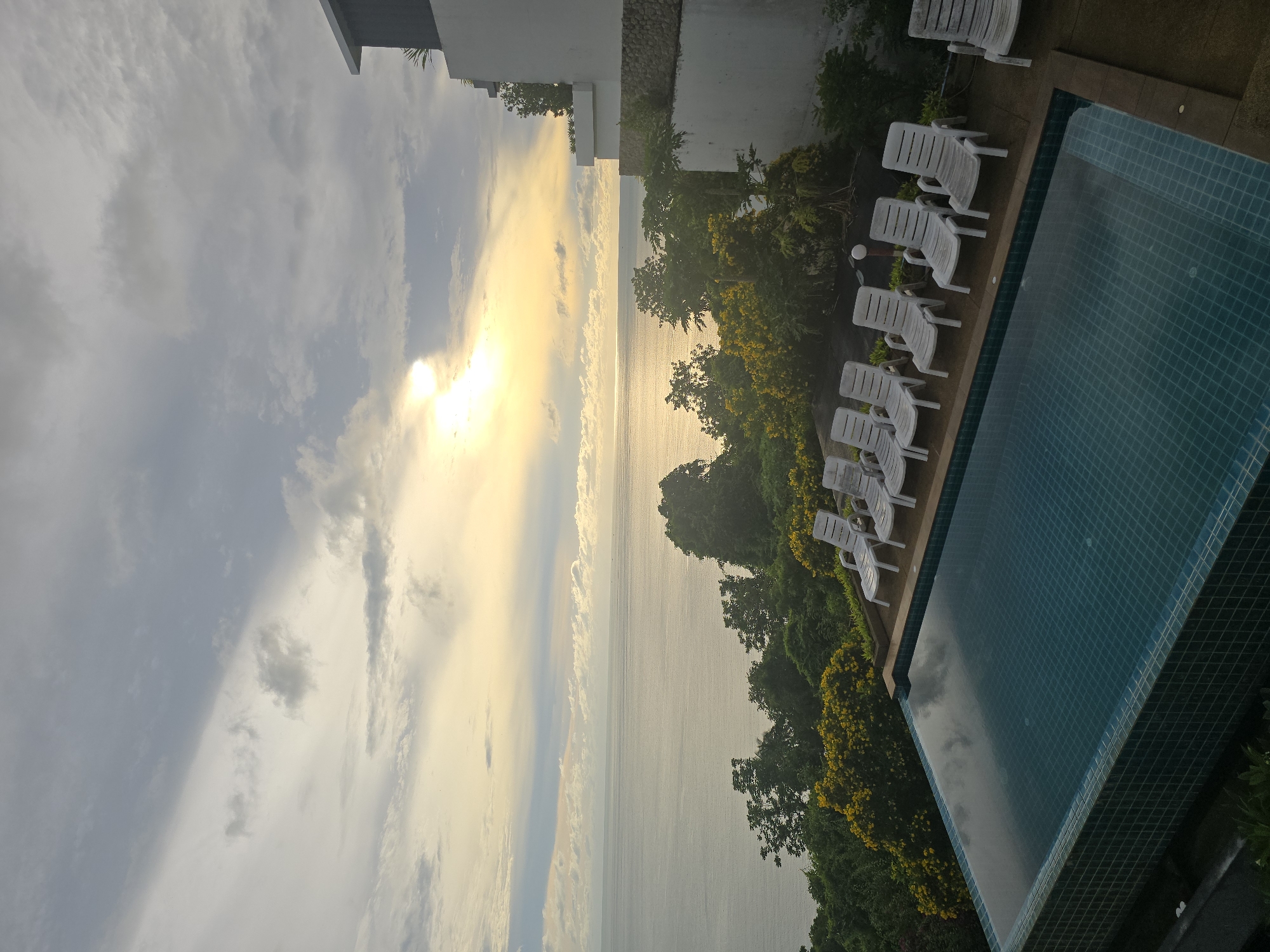 An infinity swimming pool is positioned overlooking a vast ocean and a lush, green hillside dotted with yellow flowers. The sky above is a dramatic canvas of clouds, with a bright sun peeking through, casting a golden glow on the water. Several white lounge chairs are lined up neatly beside the pool, awaiting guests. A modern building with white walls and a dark roof is visible on the right side of the frame, suggesting a resort or hotel. The overall scene conveys a sense of serene luxury and natural beauty.