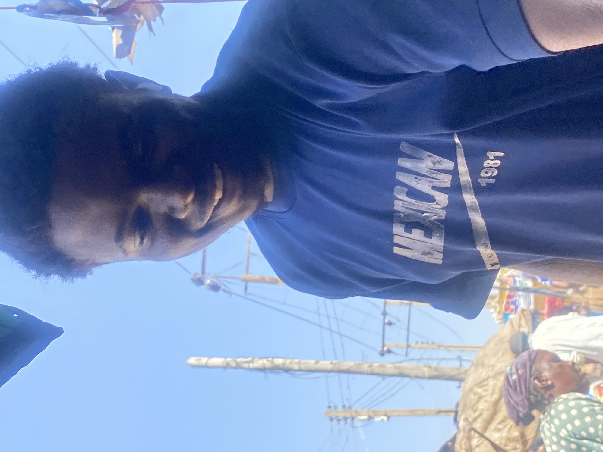 In Bukuru, Nigeria, a young Black man with dark curly hair smiles broadly, his face angled downwards and to the right. He wears a dark blue t-shirt with "MEXICAN 1981" printed in a lighter color on the front. The man is positioned in the foreground, his right shoulder and chest filling the right side of the frame. Behind him, the scene unfolds outdoors under a bright blue sky. Several utility poles with tangled wires are visible, suggesting an urban or semi-urban environment. In the mid-ground, people are seen behind a covered market stall. One woman in the lower left wears a green and white polka dot shirt and a dark headscarf. Another person, appearing to be an elderly woman, is partially visible next to her, wearing a dark purple head covering. The general atmosphere suggests a busy market day.