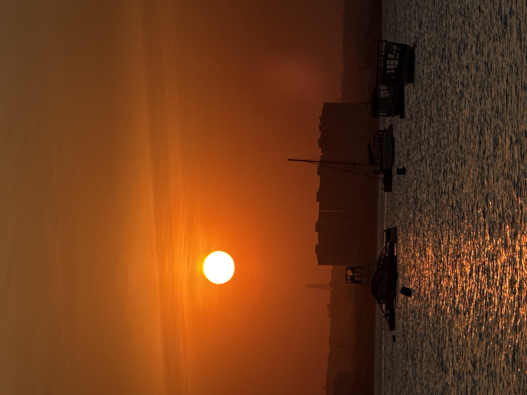 The image captures a serene sunset over the Indian Ocean, with the city of Dar es Salaam, Tanzania, silhouetted in the background. The sky is ablaze with a vibrant orange hue, dominated by a large, bright sun positioned in the upper left quadrant. Beneath the sky, the dark silhouettes of buildings and a distant tower hint at the urban landscape.
The foreground is filled with the calm, rippling waters of the ocean, reflecting the fiery colors of the sunset. Several boats are scattered across the water. In the mid-ground, a large, partially submerged structure, possibly a derelict boat or platform, sits low in the water. To its right, a modern catamaran with its sails down is visible, with what appears to be a small buoy in front of it. Further to the right, two other boats, one with a canopy and another with an open deck, are anchored. The overall scene evokes a sense of tranquility and the end of a day in this coastal East African city.