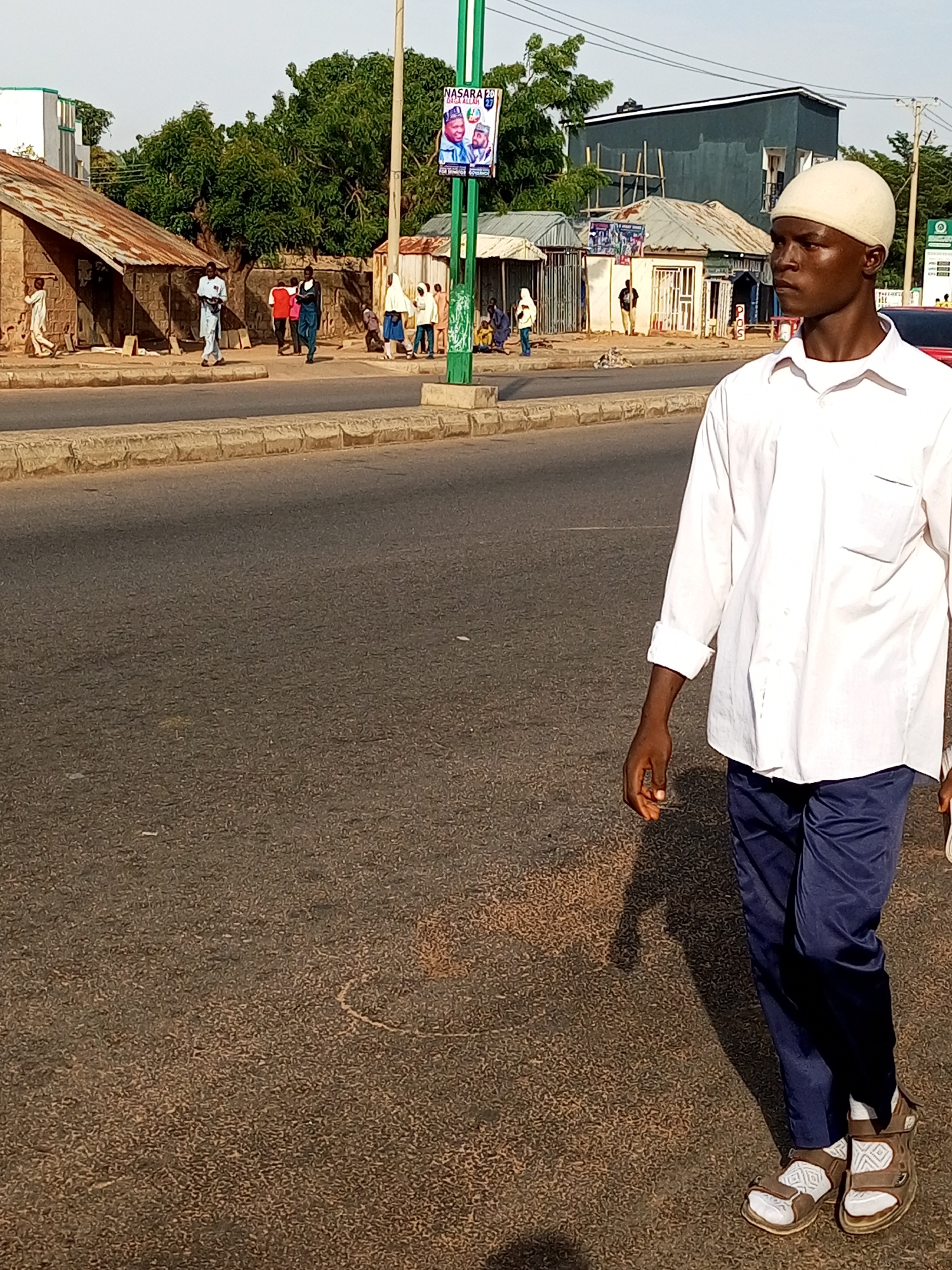 A young man wearing a white skullcap, a white long-sleeved shirt with rolled-up sleeves, and dark blue pants walks on a paved road in Garko, Nigeria. He wears white socks and brown sandals. The road has a concrete curb and a shadowed area to his left. In the background, several buildings with corrugated metal roofs and brick walls are visible. A green pole with a political poster displaying images of men and text stands near the buildings. Other people are seen walking and standing in the background. The sky is clear and the scene appears to be during daylight.