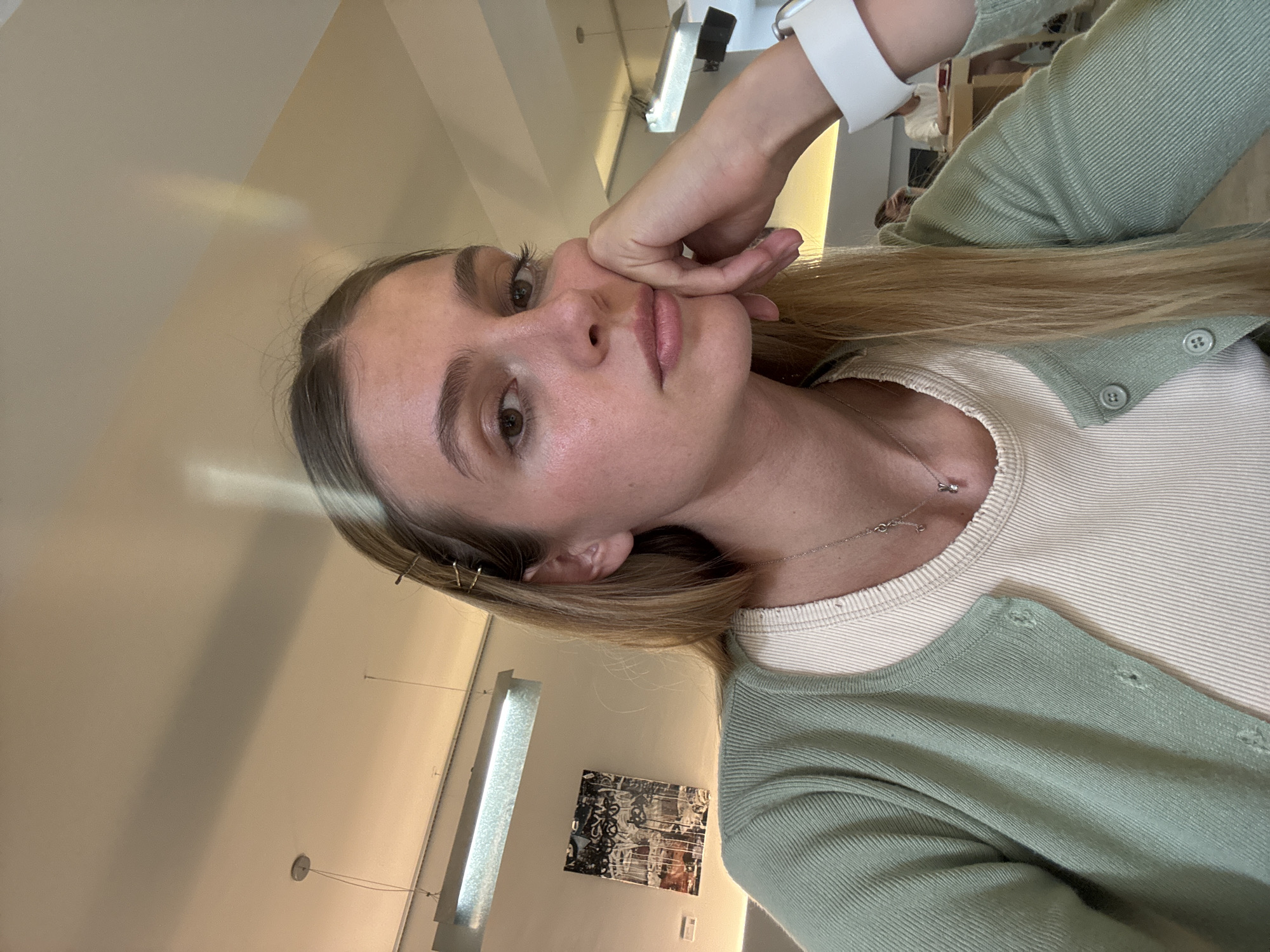 A woman with long blonde hair and light complexion is posing for a selfie, her face resting on her hand. She is wearing a light green cardigan over a light-colored, possibly white or cream, top. A thin silver necklace adorns her neck. The background appears to be an indoor setting, potentially an office or public space, with neutral-toned walls, modern lighting fixtures, and artwork on the wall. There are other people in the background of the image. The location in the city of Unknown, Unknown is not identifiable.