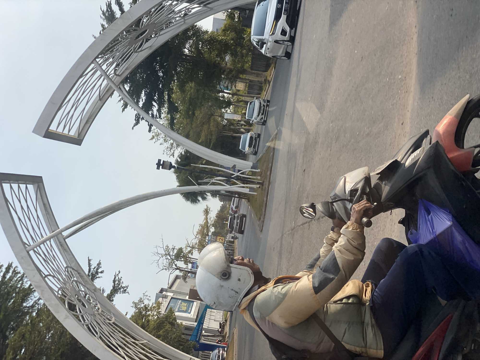 A man wearing a white helmet and a multi-colored jacket rides a motorcycle on a paved road. He is in the foreground, facing right, with his hands on the handlebars. A purple plastic bag is visible on the front of the motorcycle. The road is flanked by trees and buildings on the left, and a modern white archway structure with intricate metalwork on both sides. Several cars are visible on the road in the background, suggesting an urban or suburban environment. The sky is clear and light. The location is identified as Mandar Sari, Indonesia, with a sign visible on the left indicating "MARKETING GALLERY".