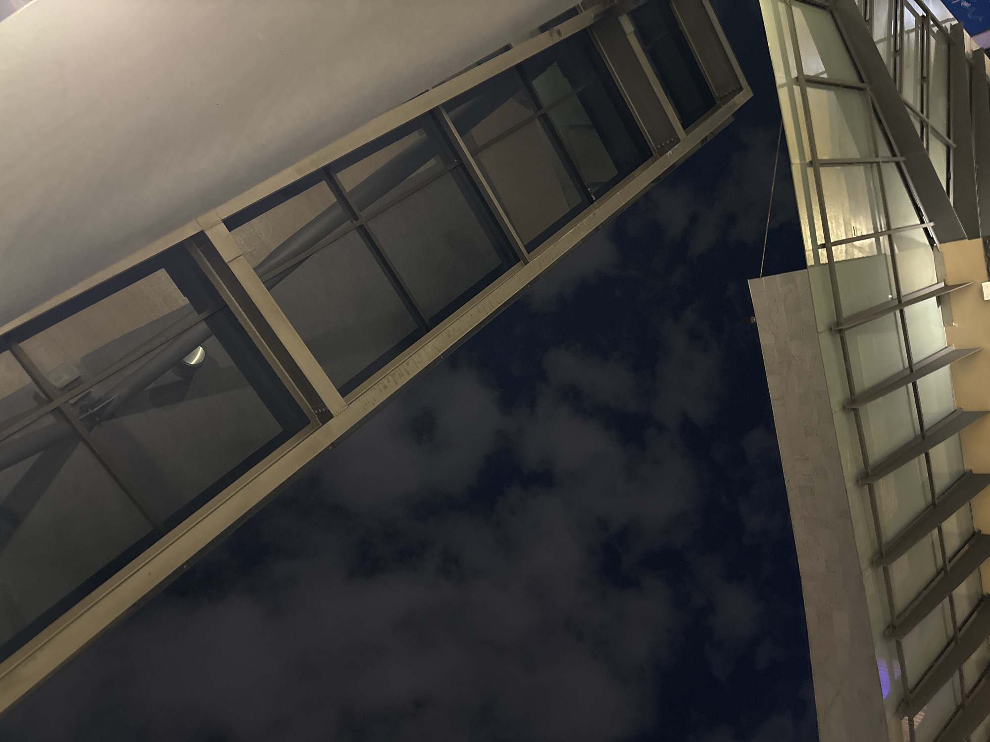 The image presents a low-angle view of modern architecture against a night sky. Two distinct structures are visible. On the left, a building facade features a grid of rectangular windows, possibly a skybridge or covered walkway. Its frame is metallic. The structure on the right is also modern, with vertical lines and repeated horizontal elements like fins or louvers. The night sky is dark with patches of visible clouds. The location is possibly in an urban area in Puebla, Mexico.