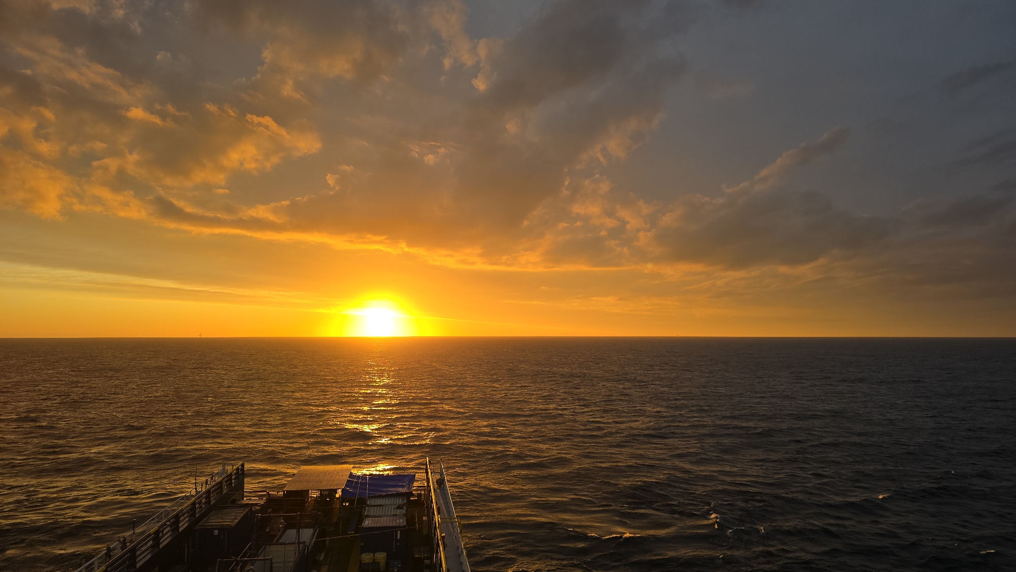 The image depicts a sunset over the ocean, viewed from what appears to be a vessel. The sun is setting on the horizon, casting a bright golden light across the sky and reflecting on the water's surface. The sky is filled with clouds, varying in color from orange to gray. The ocean is dark, with waves catching the sunlight. In the foreground, a portion of the vessel is visible, including some structures and equipment. The location is the ocean.
