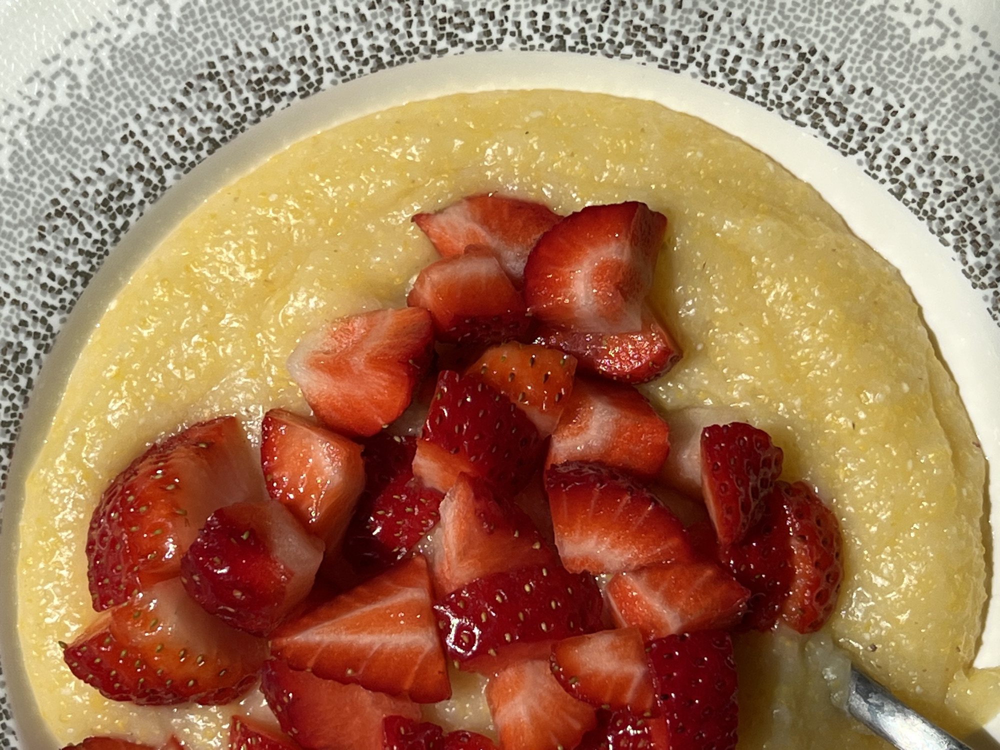 A close-up shows a bowl of polenta topped with sliced strawberries. The polenta is a smooth, yellow color, and the strawberries are bright red. The bowl is patterned with a repeating design of small gray squares. A spoon is visible in the lower right corner of the frame. The location is not discernable from this image alone. Therefore, it cannot be determined that the image is located in Palilula Urban Municipality, Serbia.