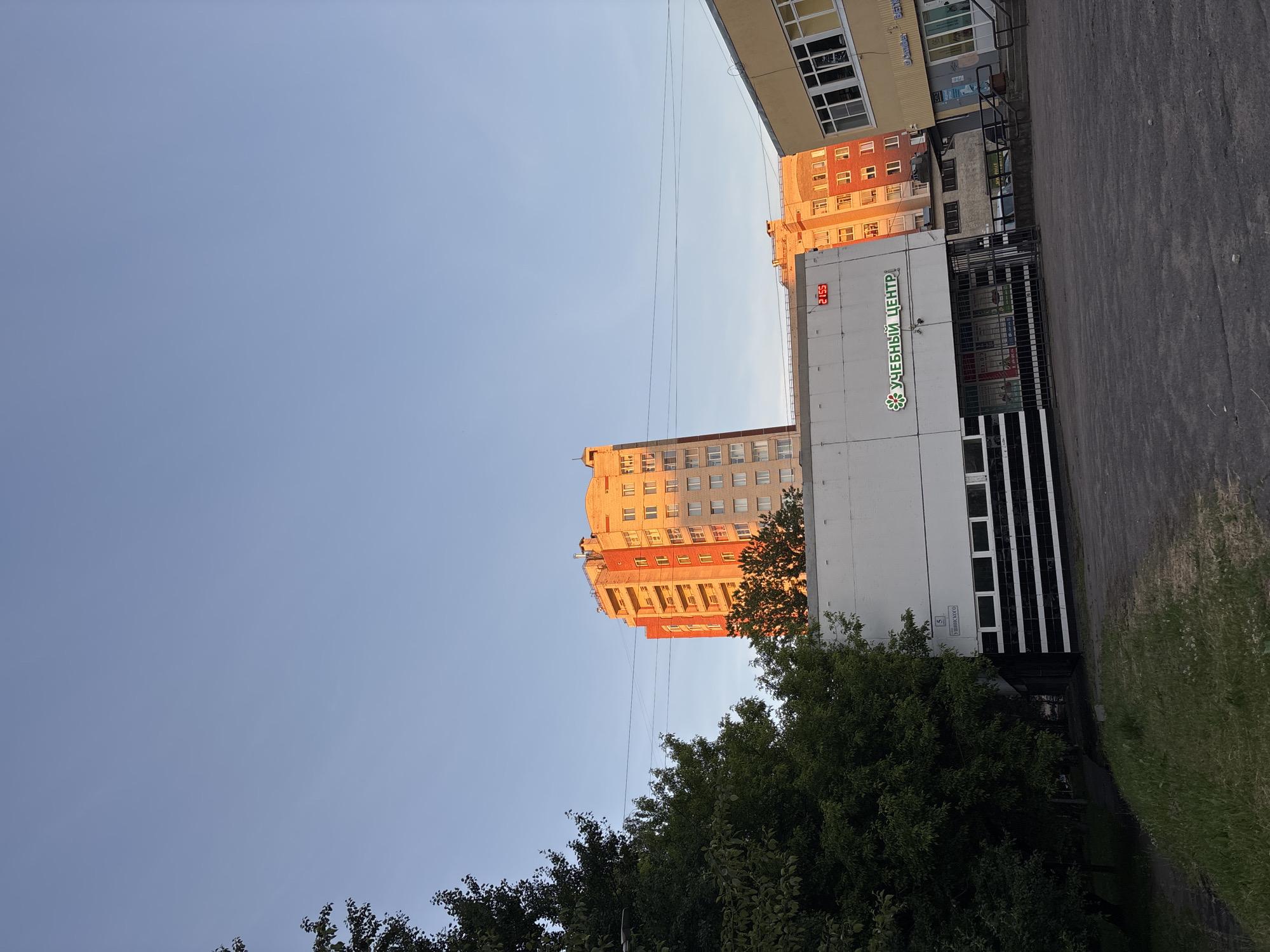 The image shows a street scene in Sinyavino, Russia. In the background, there are several tall apartment buildings with orange and beige facades, some of which are illuminated by the setting sun. In the foreground, a low-rise building with white walls and black striped garage doors is visible. A sign on this building reads "УЧЕБНЫЙ ЦЕНТР" (Educational Center) in green lettering. The sky is a clear blue, indicating it is late afternoon or early evening. There are trees on the left side of the image, casting shadows on the ground. The pavement in front of the buildings appears to be asphalt. No people are visible in the image.