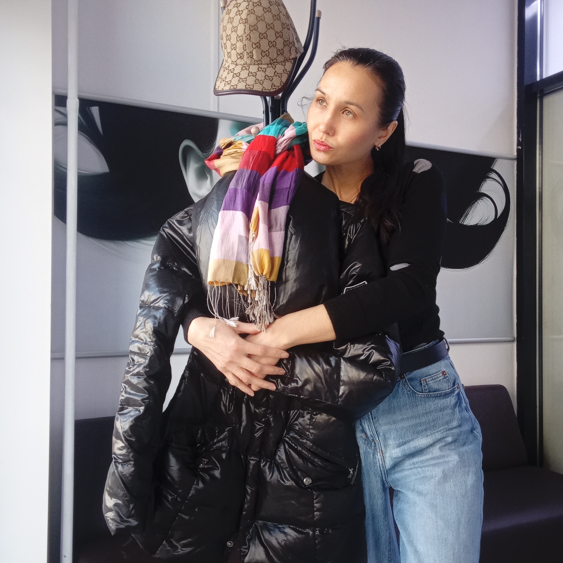 An adult woman is captured in an indoor setting, likely a modern waiting area or retail space, as suggested by the contemporary background and coat rack. She has dark hair pulled back, light-colored eyes, and a fair complexion. Her expression is thoughtful and serene as she gazes upwards and to her right. She is dressed in a black long-sleeved top, light blue denim jeans, and small silver-colored hoop earrings.
The woman is holding a shiny black puffer jacket with her hands clasped around it. A vibrant, multi-striped scarf with tassels, featuring shades of red, purple, pink, yellow, blue, and cream, is draped over the jacket and a dark-colored coat rack positioned behind her. A beige baseball cap, adorned with a repeating "GG" logo (Gucci), hangs on the top of the coat rack.
The background consists of a large, black and white graphic on a wall, displaying abstract shapes that resemble stylized human features like an ear and an eye. A plain white wall is visible to the left, and a dark window frame and part of a dark sofa or bench are on the right. The bright ambient lighting suggests it is daytime. The presence of the puffer jacket and scarf indicates cool weather. The photograph appears to be a posed shot, conveying a moment of contemplation. The location is Izhevsk, Russia.
