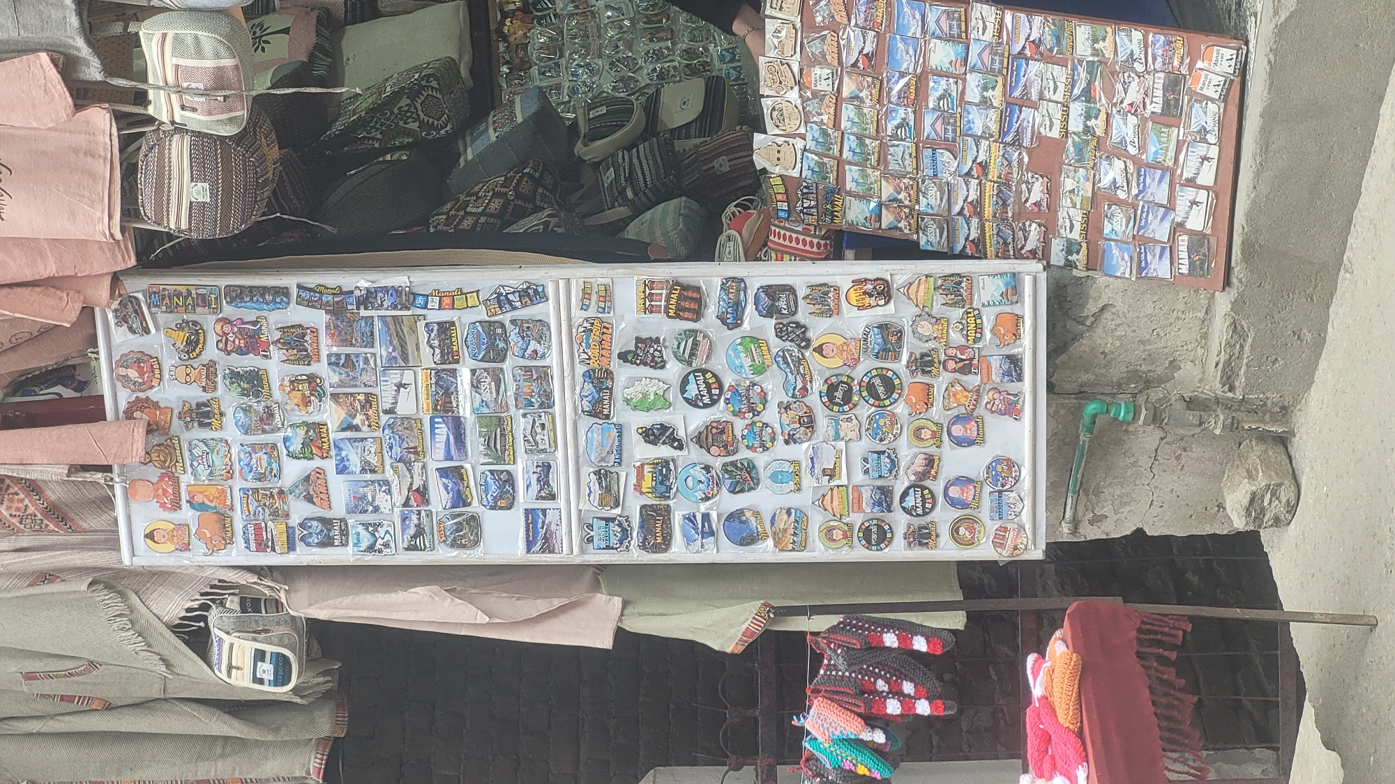 The image depicts a street stall in Vashisht, India, selling various souvenirs. The stall features a large display of colorful stickers and magnets, many of which appear to be related to local tourism, with text such as "Manali" and "Sissu" visible. These are arranged on a white board and a brown board. To the left of the sticker display, there are knitted items, possibly hats or bags, hanging and displayed on a red cloth-covered table. Behind the stall, traditional textiles are hung, and bags are displayed, suggesting a marketplace environment. A green pipe and a concrete wall are also visible in the foreground and background, indicating an outdoor setting. The overall scene suggests a vibrant, commercial area catering to tourists.