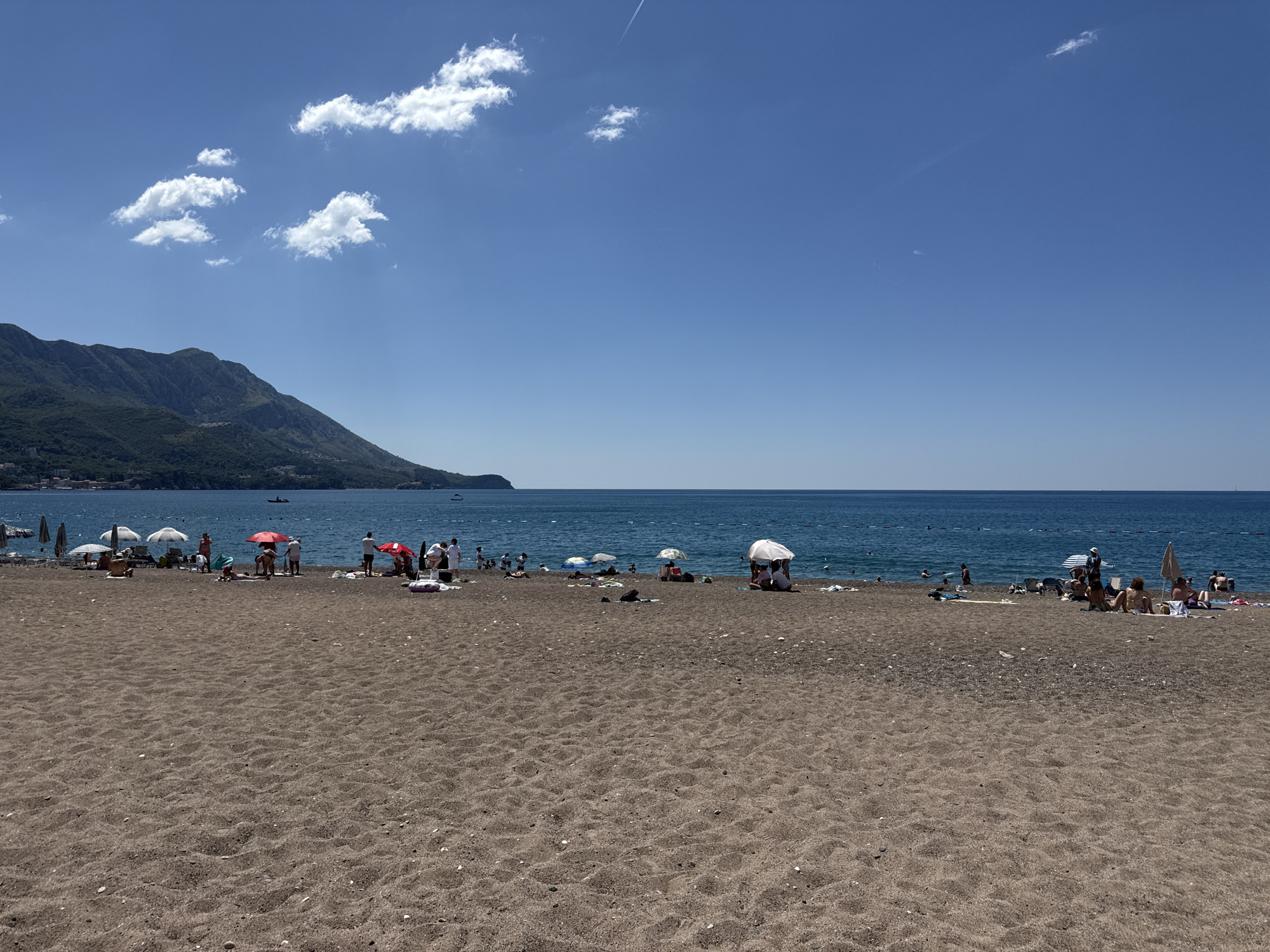 This image captures a sunny day at the beach in Boreti, Montenegro. The foreground is dominated by a wide expanse of dark sand, with visible pebbles and shells scattered across its surface. In the middle ground, a bustling beach scene unfolds. Numerous people are spread out on the sand, some reclining under beach umbrellas, while others are standing or walking. A variety of colorful umbrellas, both open and closed, provide shade. Several individuals are in or near the water. The sea itself is a deep blue, calm and stretching towards the horizon, with a few buoys visible in the water marking a swimming area. In the background, a lush green, mountainous landscape rises from the coastline, with buildings dotting the slopes. The sky above is a clear, bright blue, with a few scattered white clouds. A faint contrail from an aircraft is visible in the upper right portion of the sky. The overall impression is one of a popular, lively beach resort on a beautiful summer day.